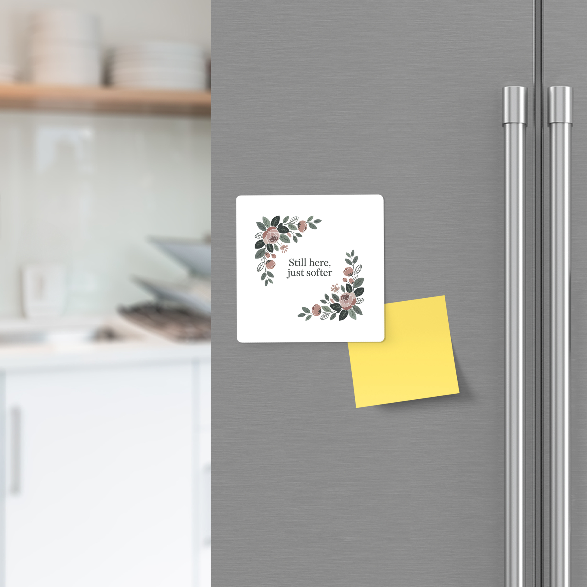 The Mirabilia Boutique Still Here Just Softer Floral Magnet, featuring a botanical design and uplifting text, is displayed on a gray refrigerator door beside a yellow sticky note—an ideal encouragement gift. Blurred kitchen shelves appear in the background.