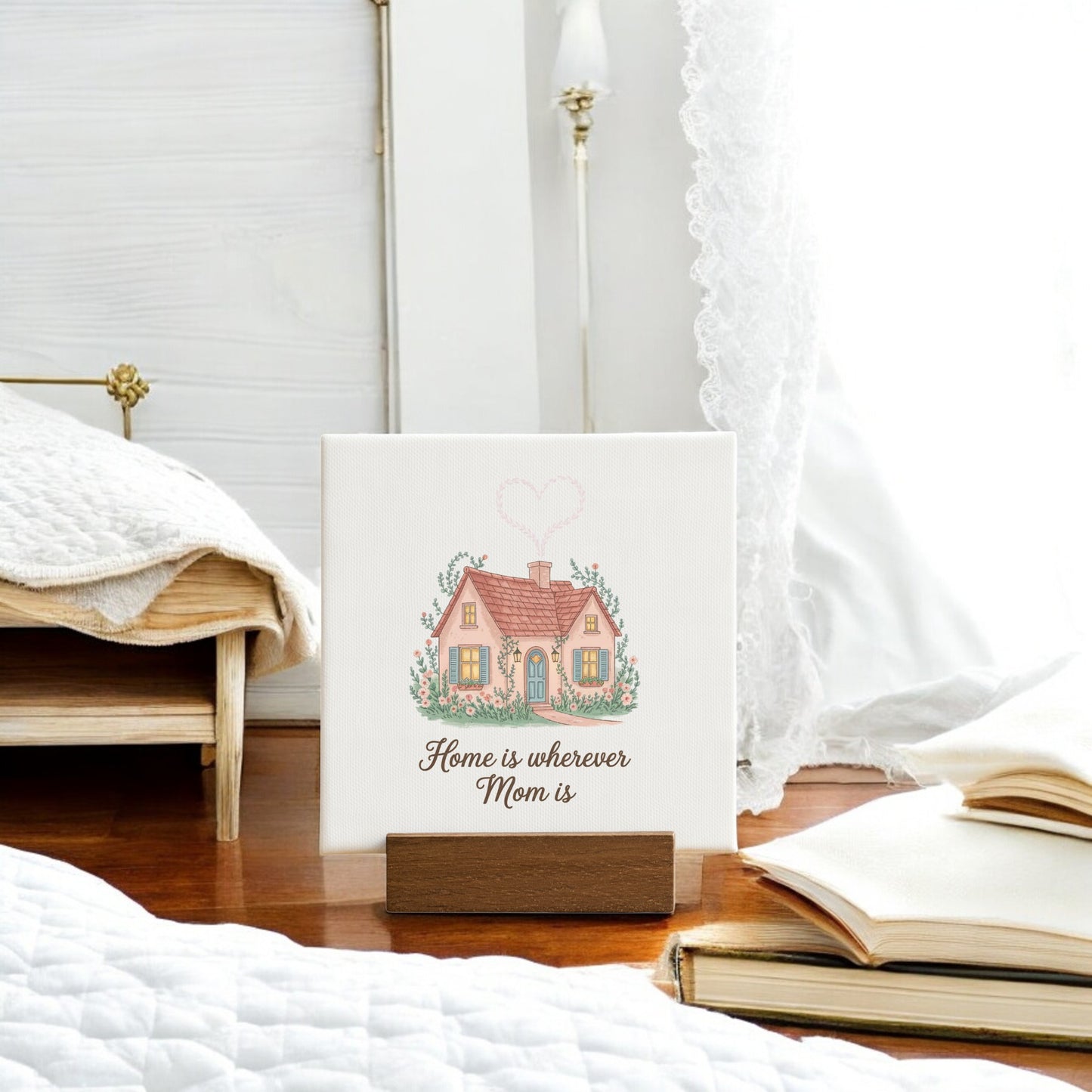 The Mirabilia Boutique "Home Is Wherever Mom Is" Mini Sign features a house design on a wooden stand—ideal as a Mother’s Day gift, it looks charming in a cozy, white bedroom beside an open book.