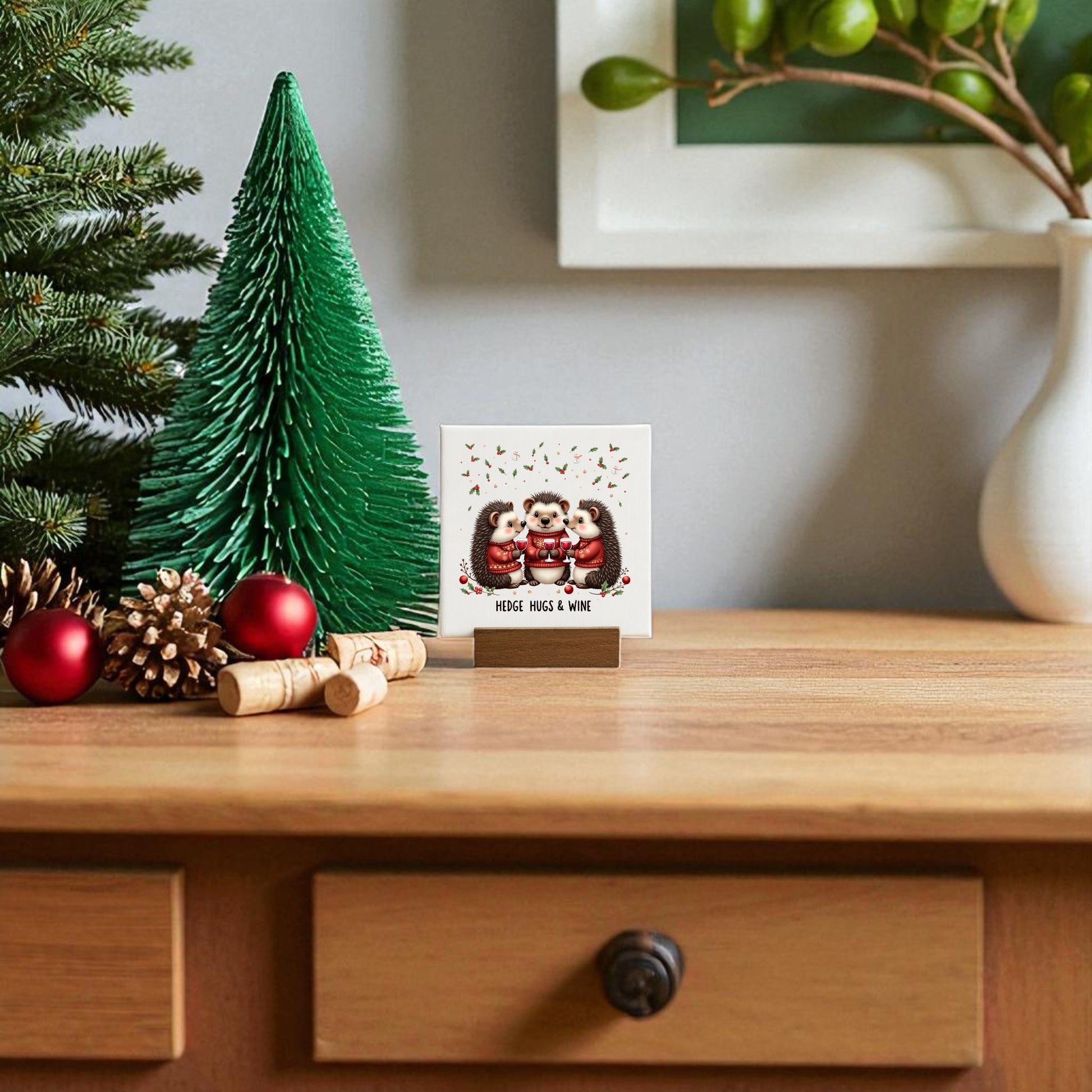 A festive wooden table features pine branches, red ornaments, pinecones, a green paper Christmas tree, and a greeting card with three owls in winter gear. The “Hedge Hugs & Wine” canvas sign by Mirabilia Boutique adds whimsical charm; greenery in a white vase sits behind.