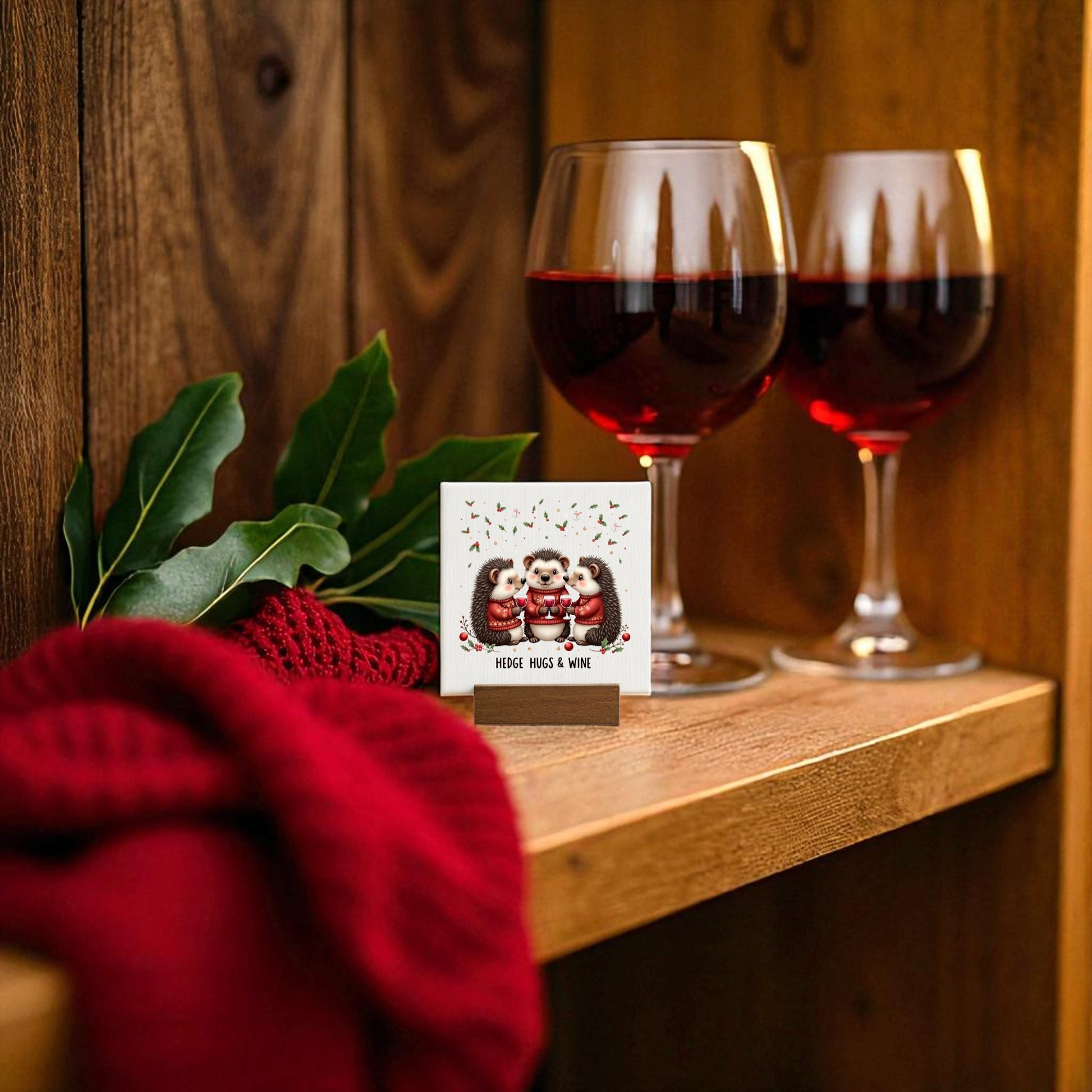 The Hedge Hugs & Wine Canvas Sign by Mirabilia Boutique features red wine glasses, a scarf, and hedgehogs with mugs—ideal as cozy holiday decor or a wine lover gift. This 4x4 sign adds charm to any space.