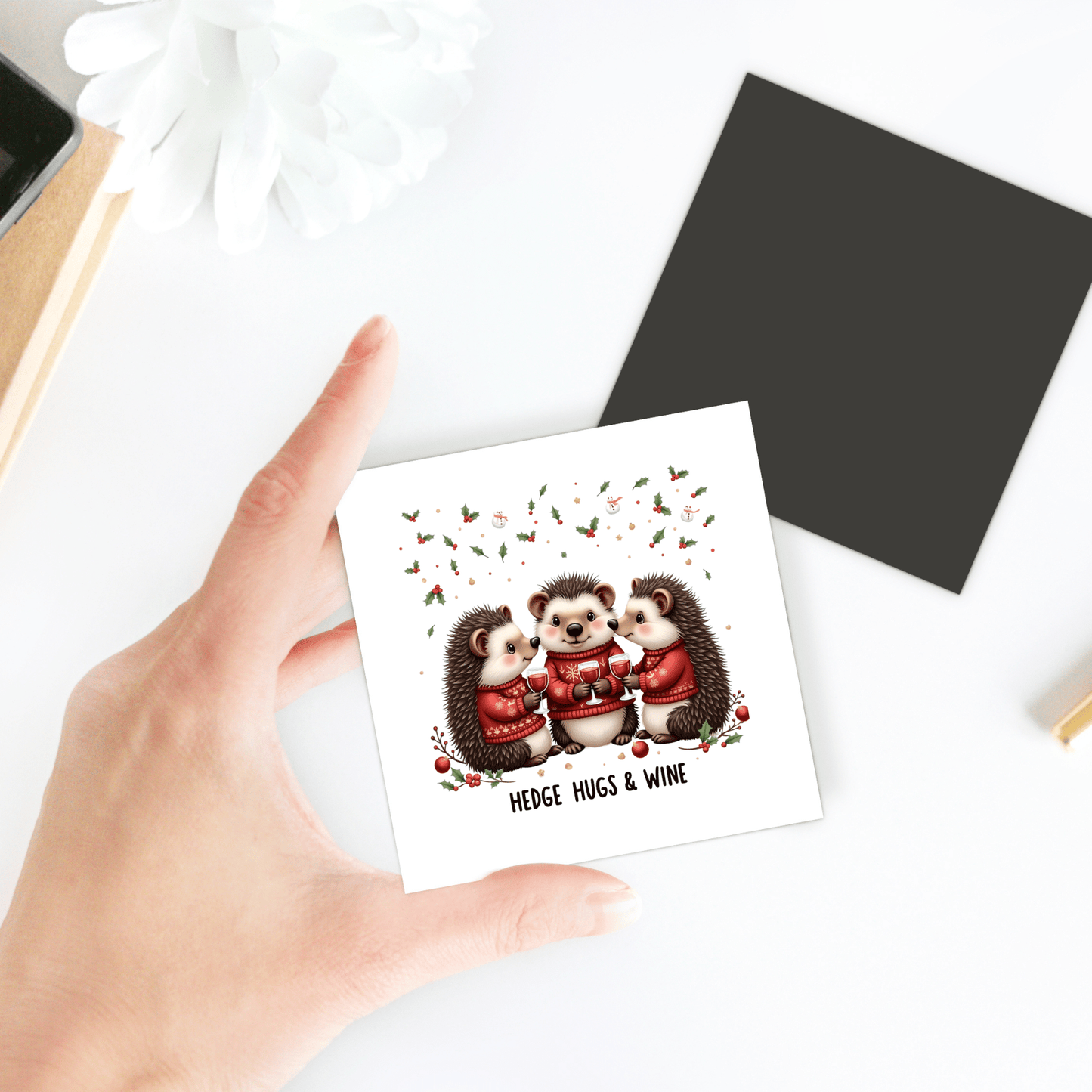 A hand holds the "Hedge Hugs & Wine Magnet" by Mirabilia Boutique, featuring three cute hedgehogs in sweaters with floral accents—a funny holiday gift for wine lovers or charming fridge decor.