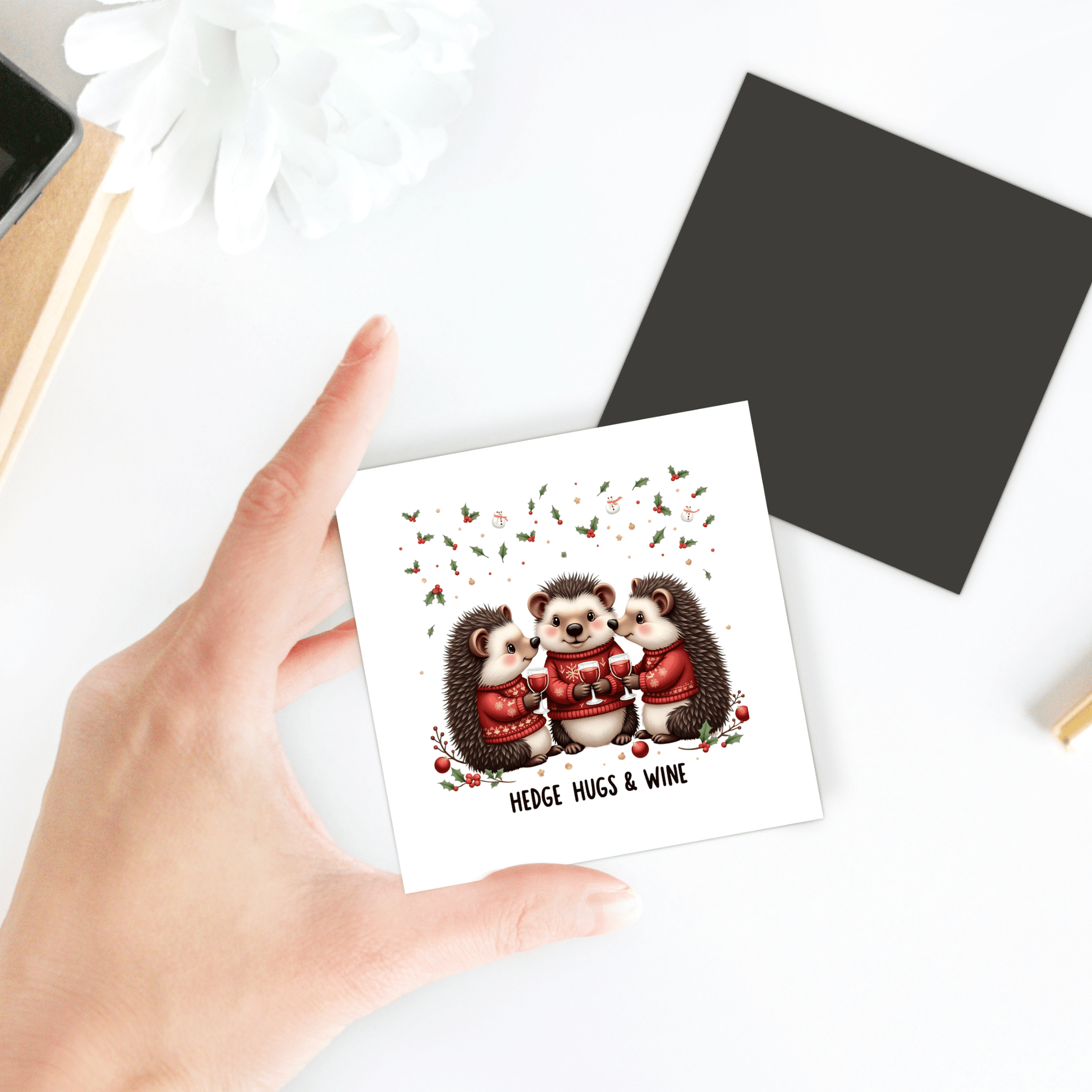 A hand holds the "Hedge Hugs & Wine Magnet" by Mirabilia Boutique, featuring three cute hedgehogs in sweaters with floral accents—a funny holiday gift for wine lovers or charming fridge decor.