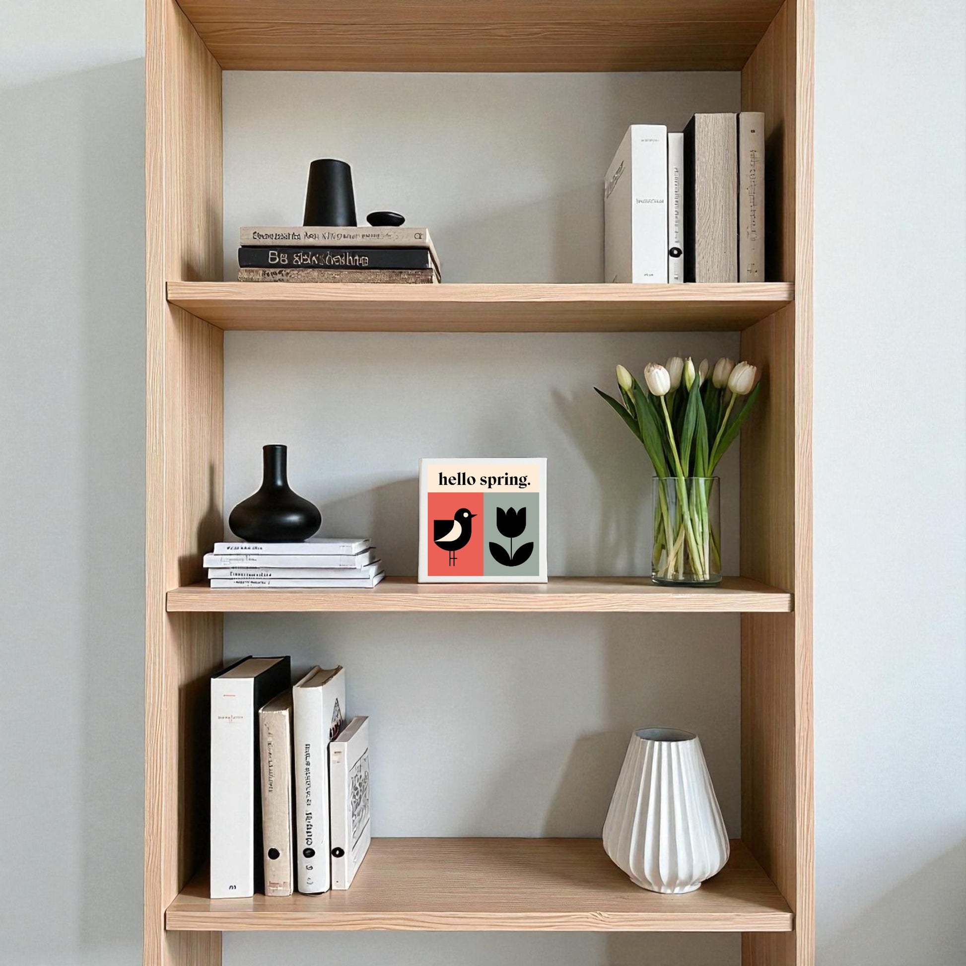 The Hello Spring Canvas – Bird & Tulip Wall Art by Mirabilia Boutique adds cheerful, modern decor to your home when paired with a wooden bookshelf, black and white vases, white tulips in glass, and neatly arranged books.