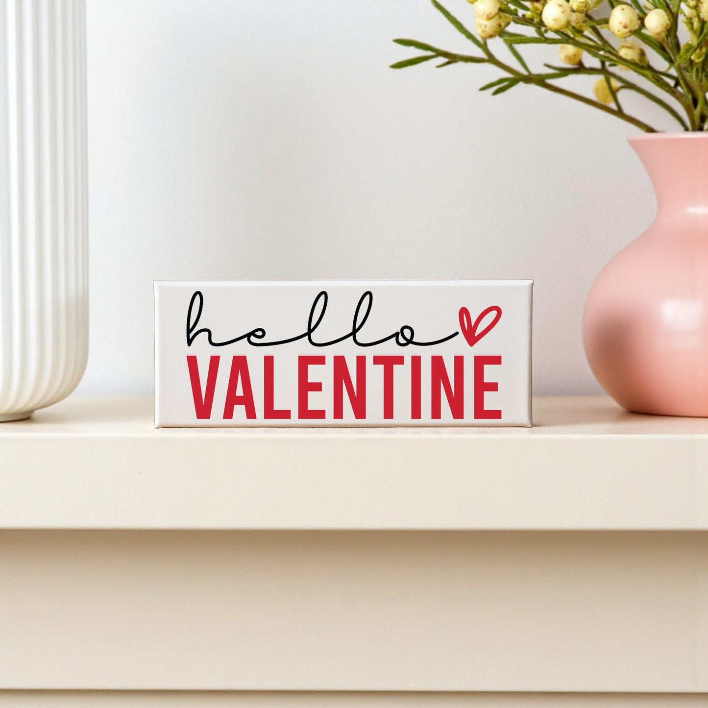 The Mirabilia Boutique Hello Valentine Sign | Modern Red & White Shelf Decor adds a charming touch to your romantic home when displayed on a white shelf beside a pink vase and decorative accents for Valentine’s Day.