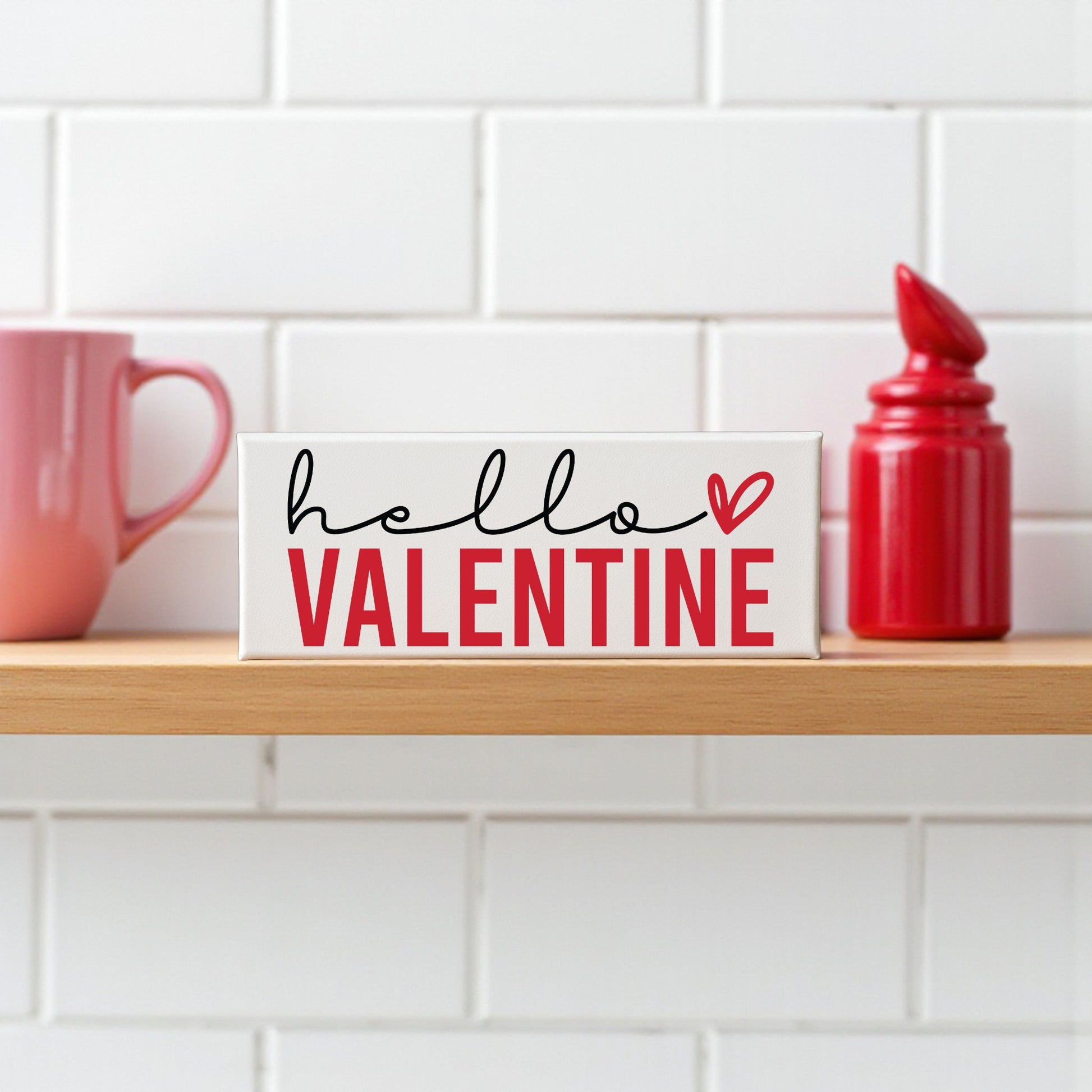 The Mirabilia Boutique Hello Valentine Sign | Modern Red & White Shelf Decor sits on a wooden shelf with a pink mug and red jar, adding charming Valentine’s Day style against a white tiled wall.