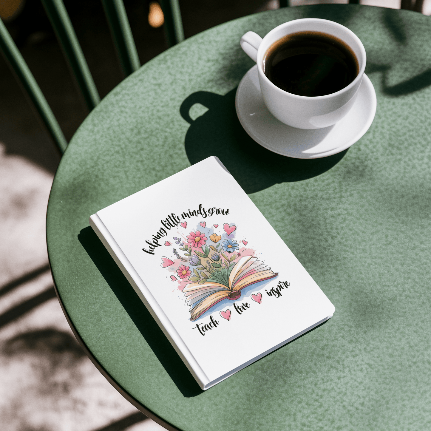 The Mirabilia Boutique “Helping Little Minds Grow” Hardcover Journal, featuring a colorful floral and book design with the phrase "Teach Love Inspire," rests on a green table beside a cup of black coffee.