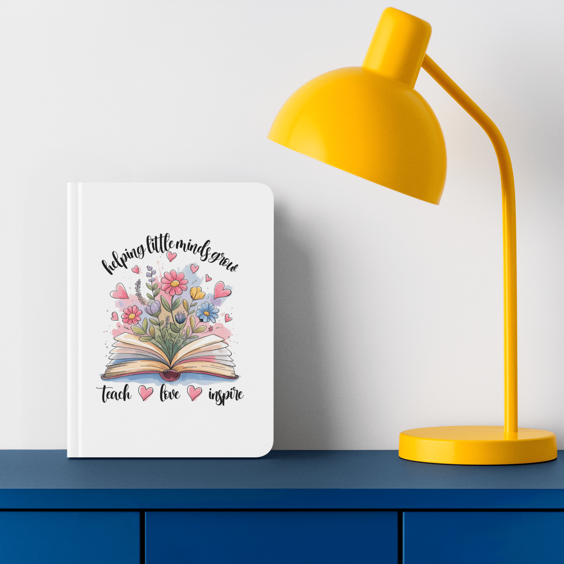 The Mirabilia Boutique "Helping Little Minds Grow" Hardcover Journal features an open book, colorful flowers, and pink hearts on its cover. This teacher gift notebook is pictured next to a yellow desk lamp on a blue surface.