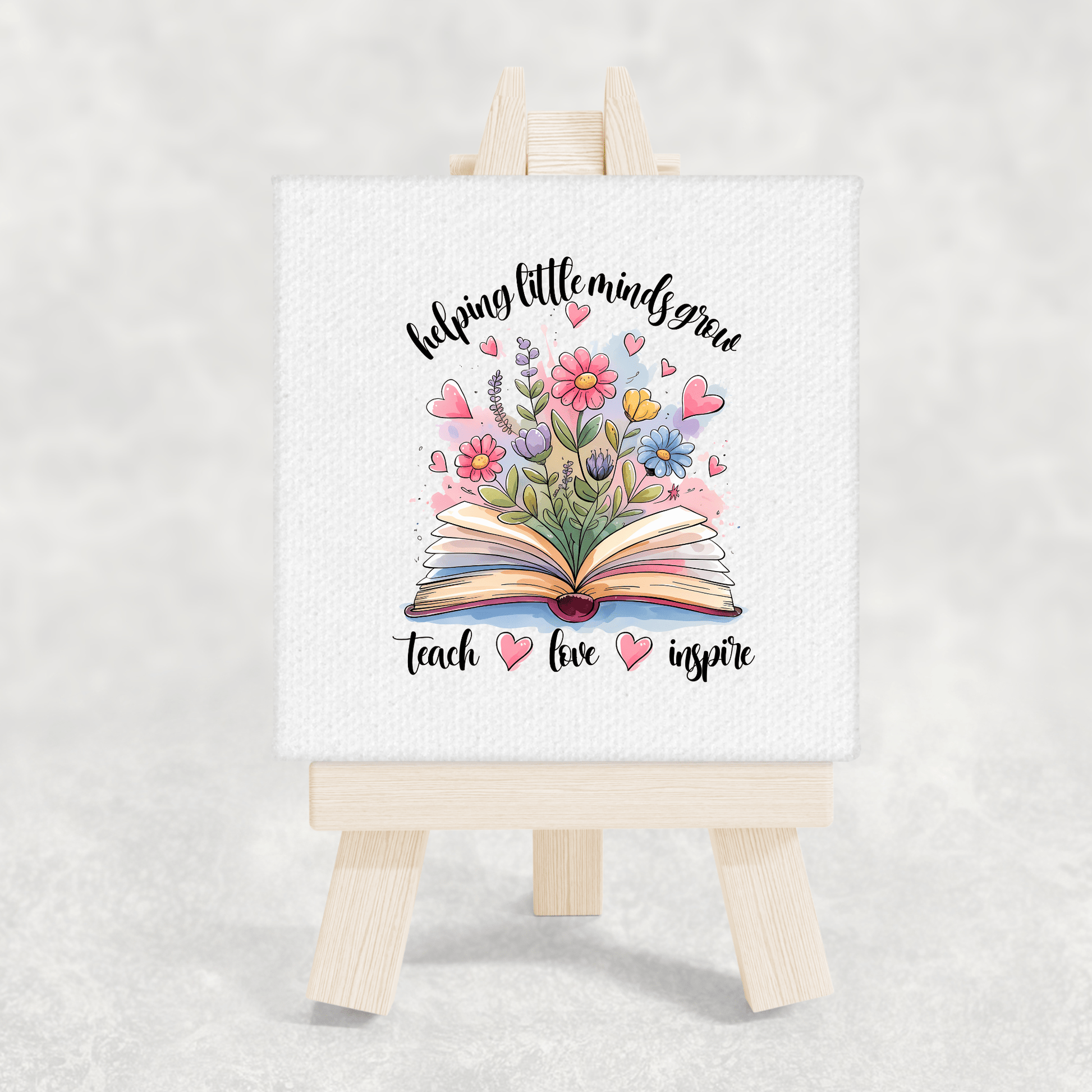 The Mirabilia Boutique Helping Little Minds Grow Mini Canvas with easel displays an open book with flowers and hearts, plus the phrase “helping little minds grow, teach, love, inspire”—a perfect teacher gift or classroom decor.