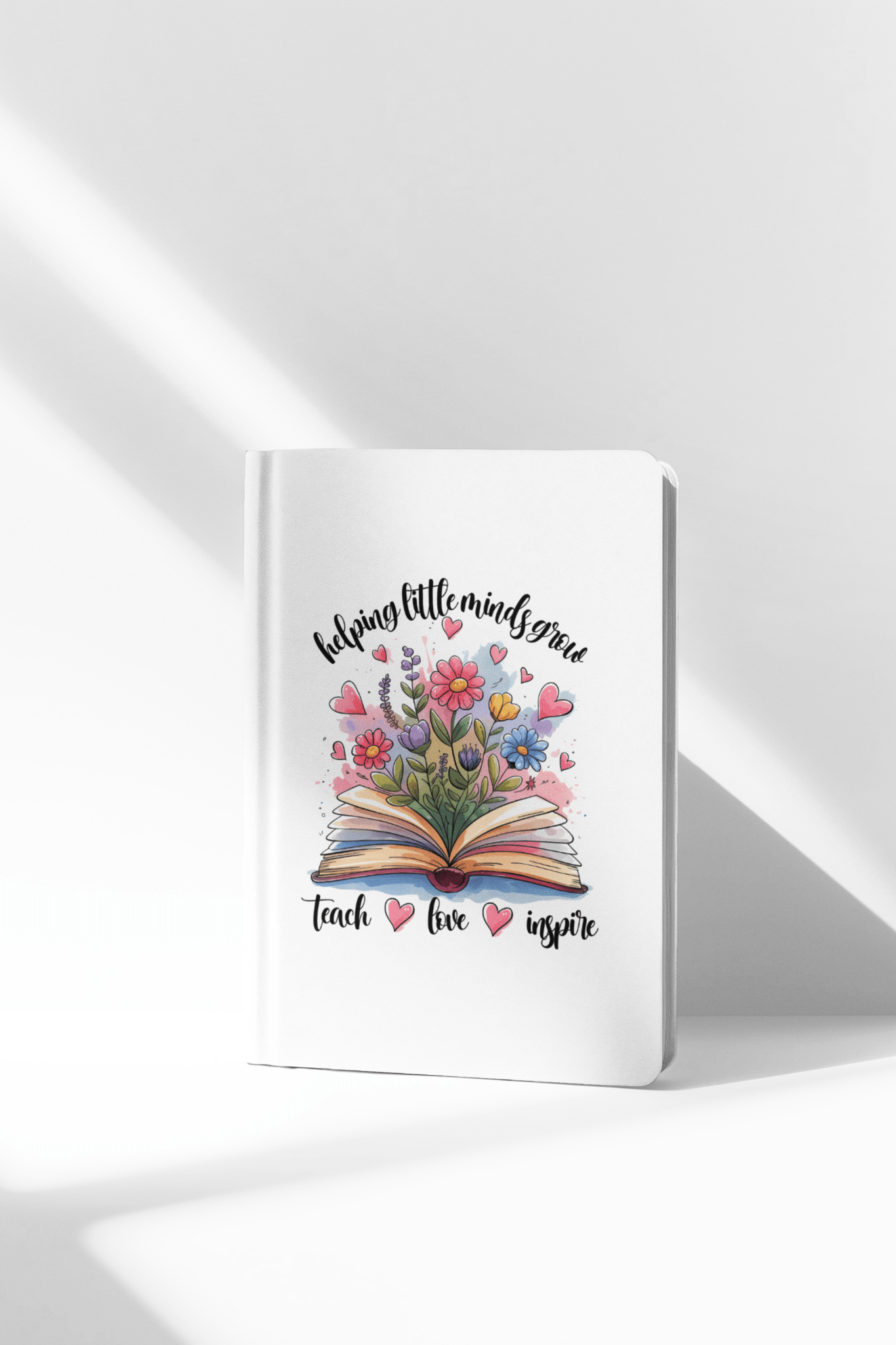 The Helping Little Minds Grow Hardcover Journal by Mirabilia Boutique features an illustrated cover with an open book, colorful flowers, and hearts. The text says, "Helping little minds grow. Teach. Love. Inspire." Ideal teacher gift or journal.