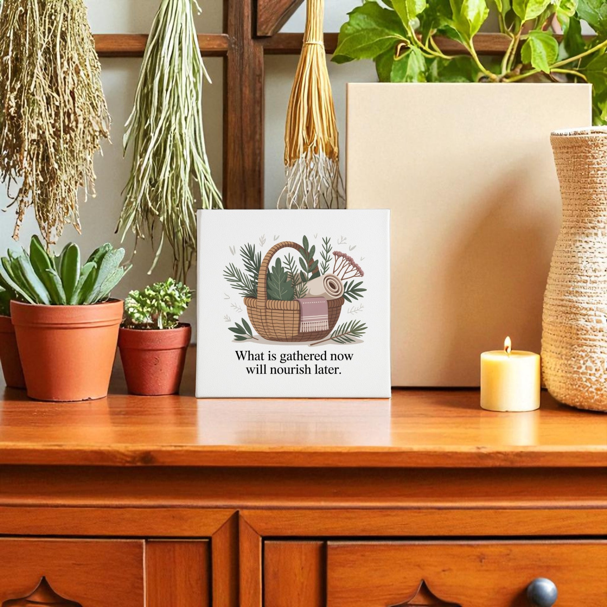 A wooden table displays potted plants, a candle, and Mirabilia Boutique’s Herbal Basket Canvas Art—an inspiring wall sign with the quote "What Is Gathered Now Will Nourish Later"—creating a cozy slow living vibe among greenery and dried herbs.