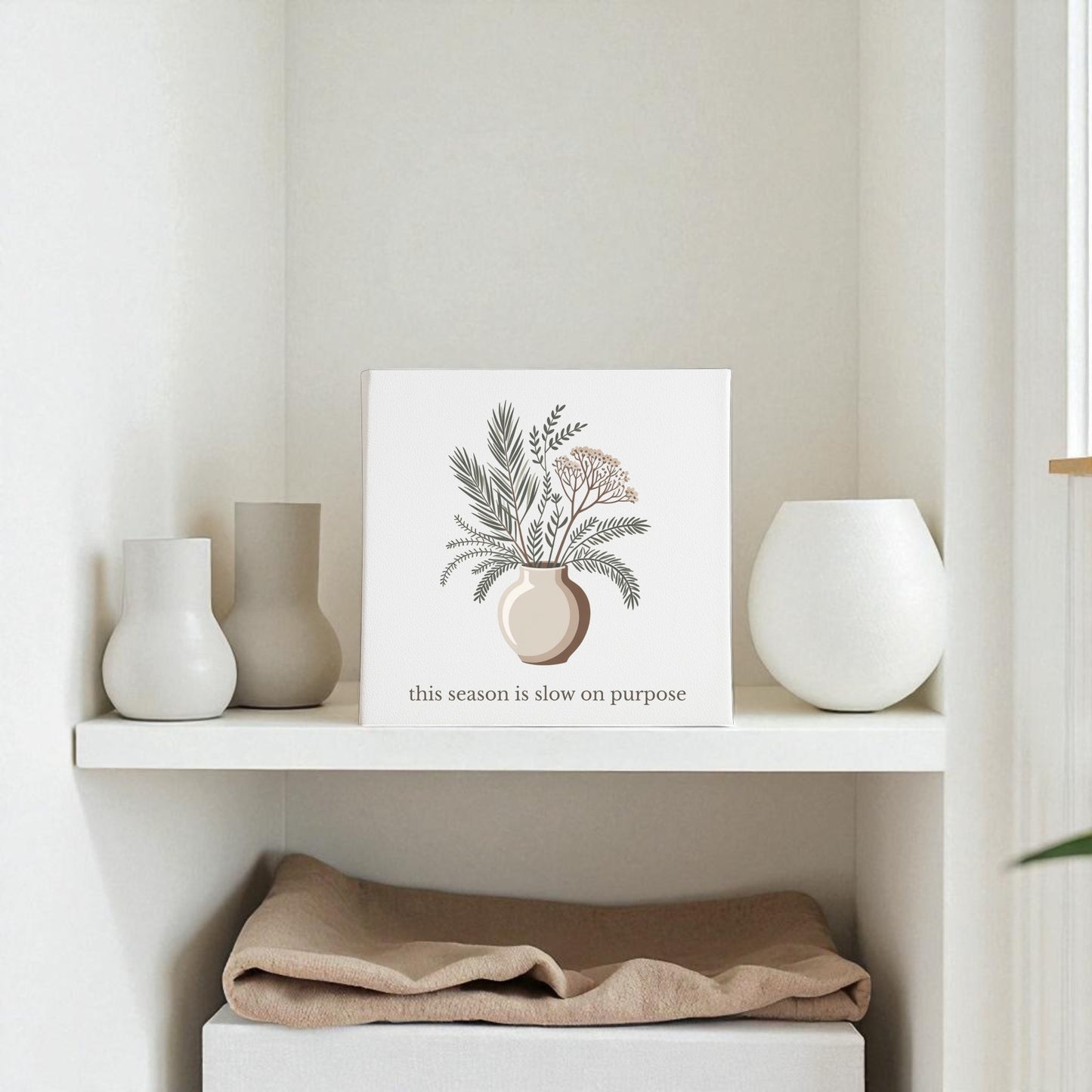 A minimalist shelf displays neutral vases and Mirabilia Boutique’s “Slow on Purpose Canvas | Botanical Vase Quote Wall Art," embracing intentional living, with a folded beige cloth on the lower shelf.