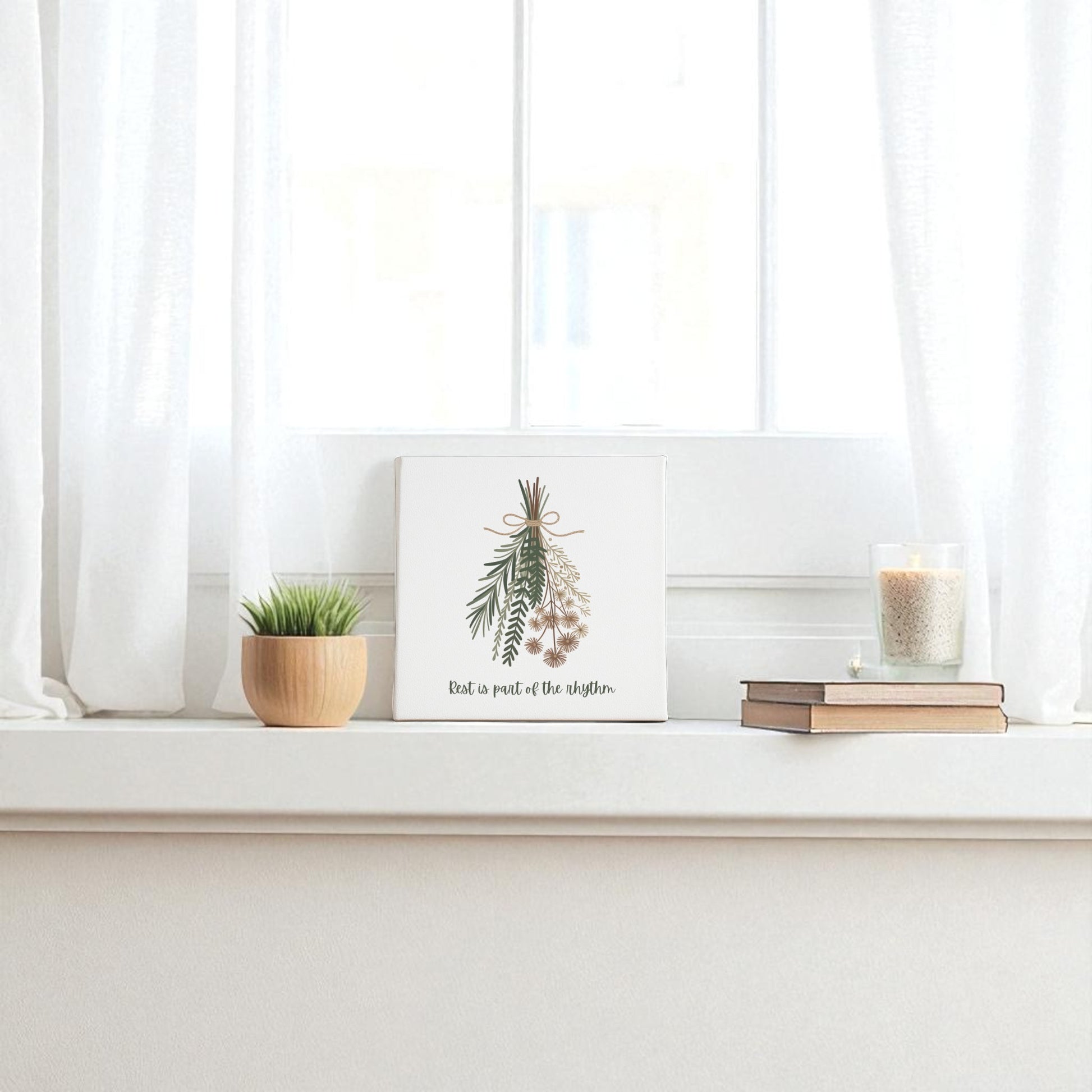 A white windowsill features a potted plant, stacked books, and a lit candle in glass. The “Rest Is Part of the Rhythm” botanical canvas by Mirabilia Boutique adds minimalist charm, while white curtains frame the bright window for seasonal harmony.