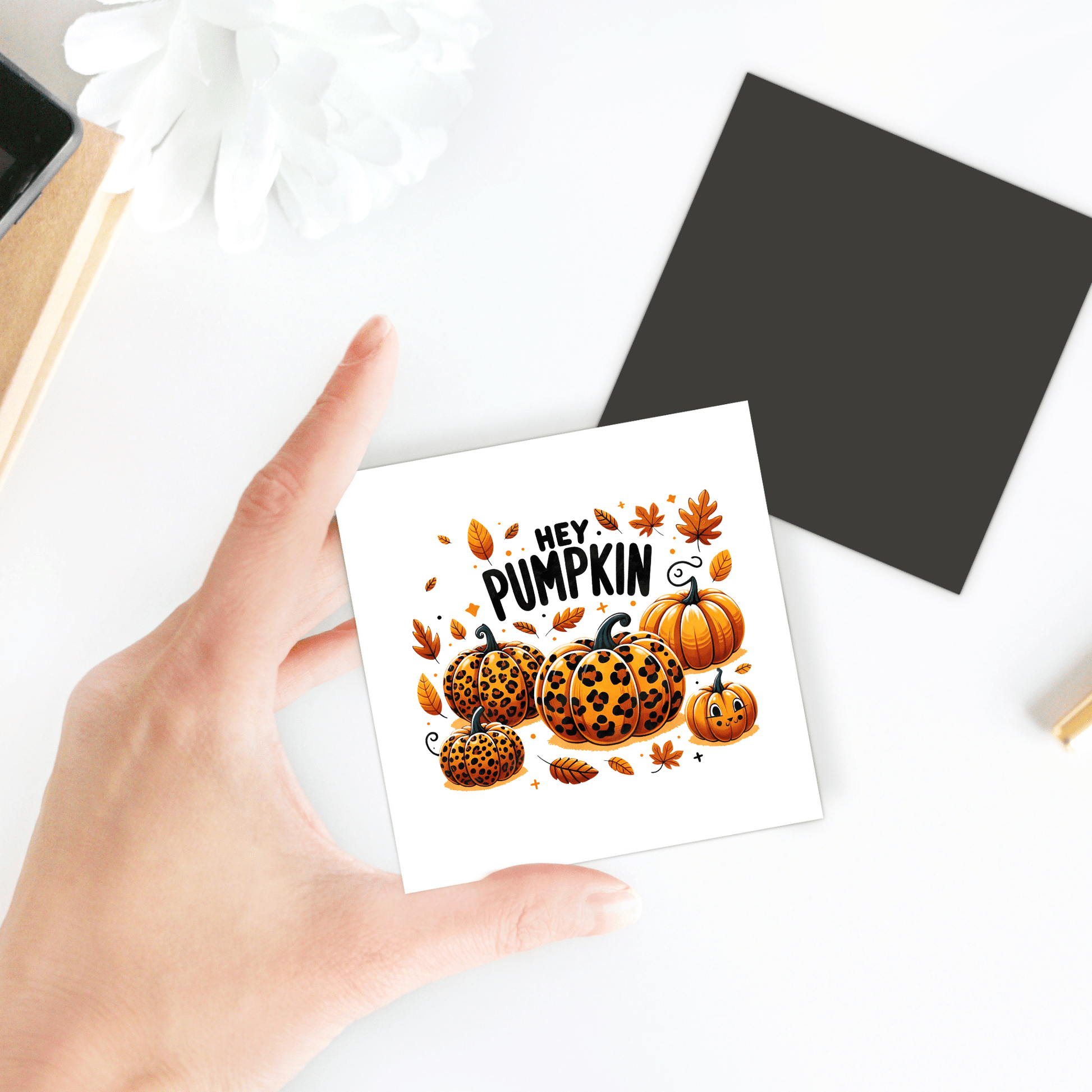 A hand displays the Mirabilia Boutique "Hey Pumpkin" Magnet—featuring orange and leopard-print pumpkins and autumn leaves—for stylish fall fridge decor. Nearby are a black square and a white flower on the table.