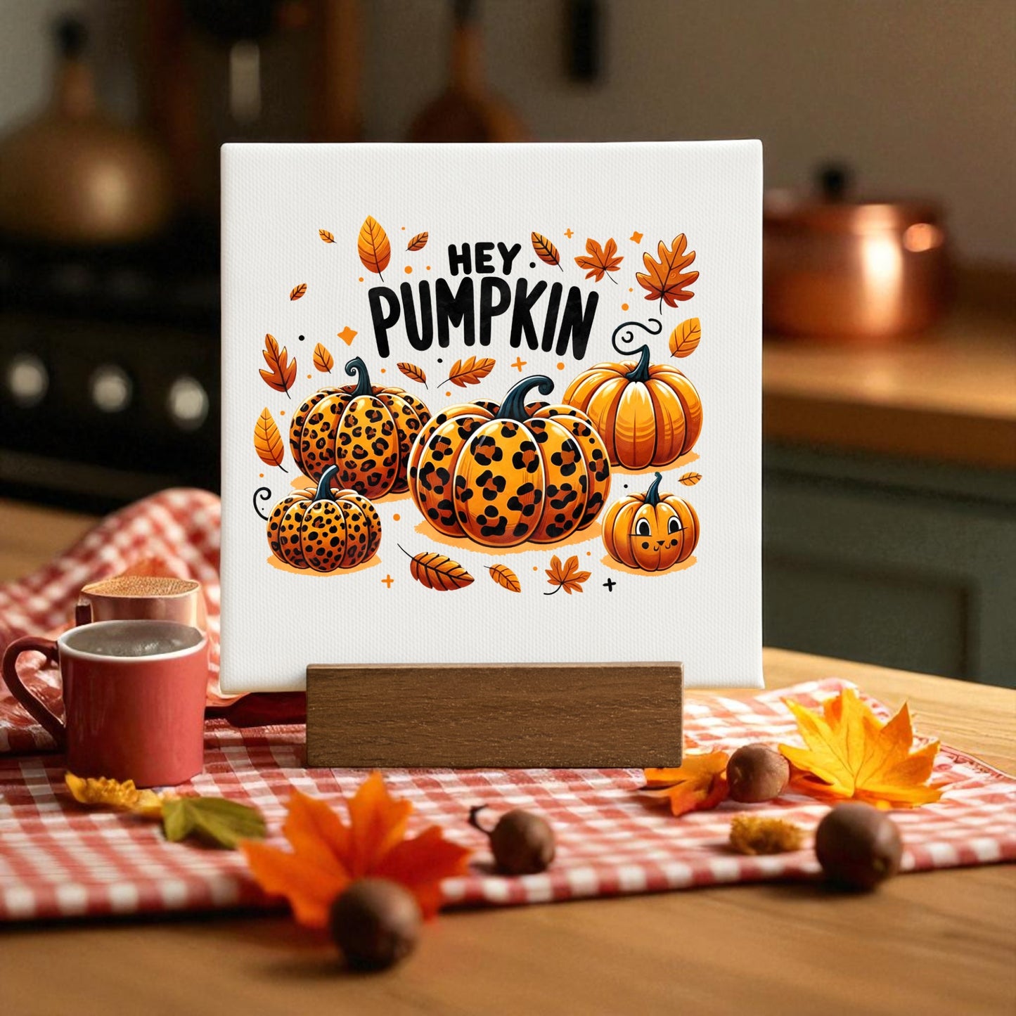 A kitchen displays the "Hey Pumpkin Mini Sign" by Mirabilia Boutique, featuring leopard print pumpkins and autumn leaves. Fall decor and a mug sit on a checkered cloth, adding charm to the cozy fall scene.