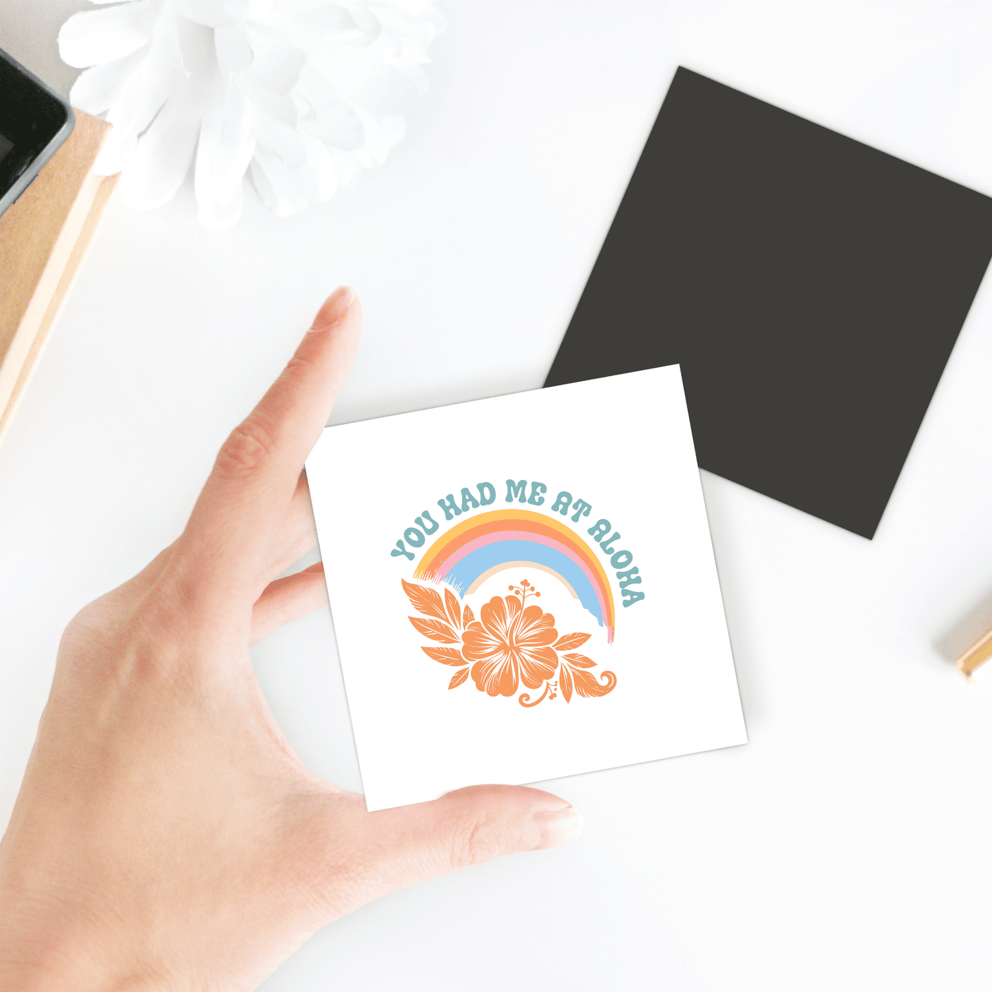 A hand holds the Mirabilia Boutique "You Had Me at Aloha Magnet"—a square magnet with a rainbow, hibiscus, and blue text, perfect for beach house decor. A black tropical fridge magnet sits nearby on the white surface.