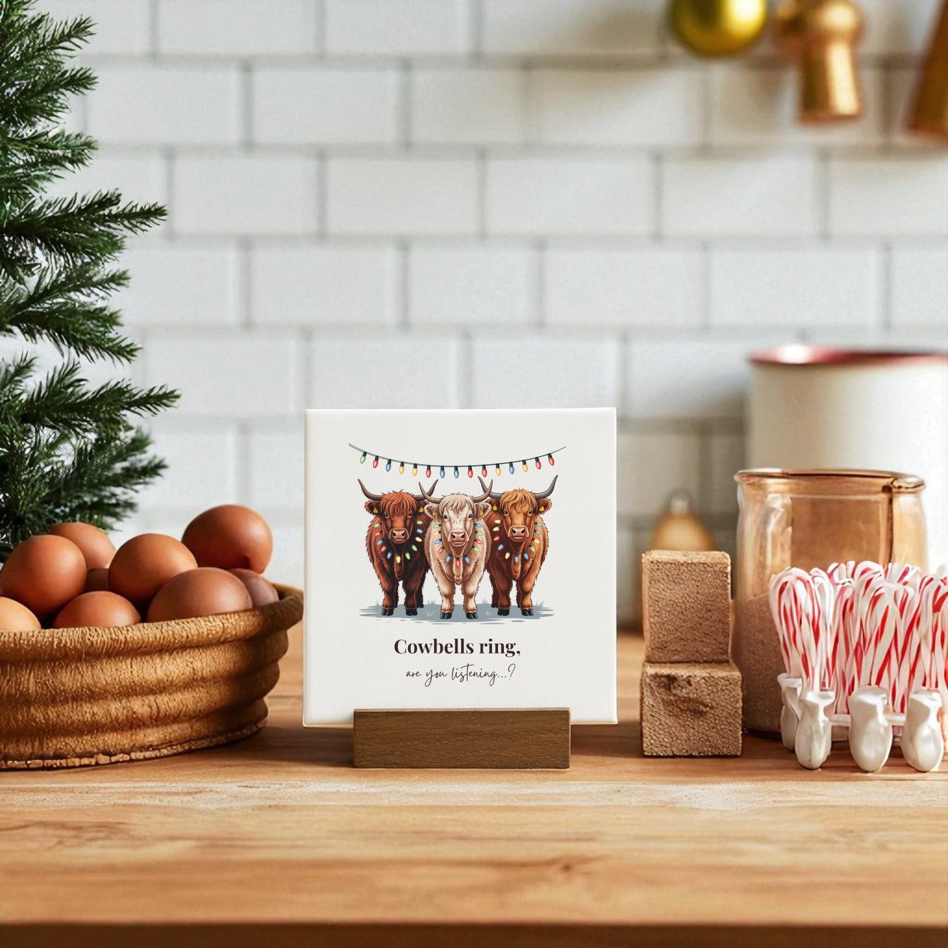 A festive kitchen scene features the Mirabilia Boutique Highland Cow Christmas Mini Sign (4x4), a rustic decor piece with three cows in holiday lights and the phrase "Cowbells ring, are you listening?", surrounded by eggs, candy canes, and fir branches.