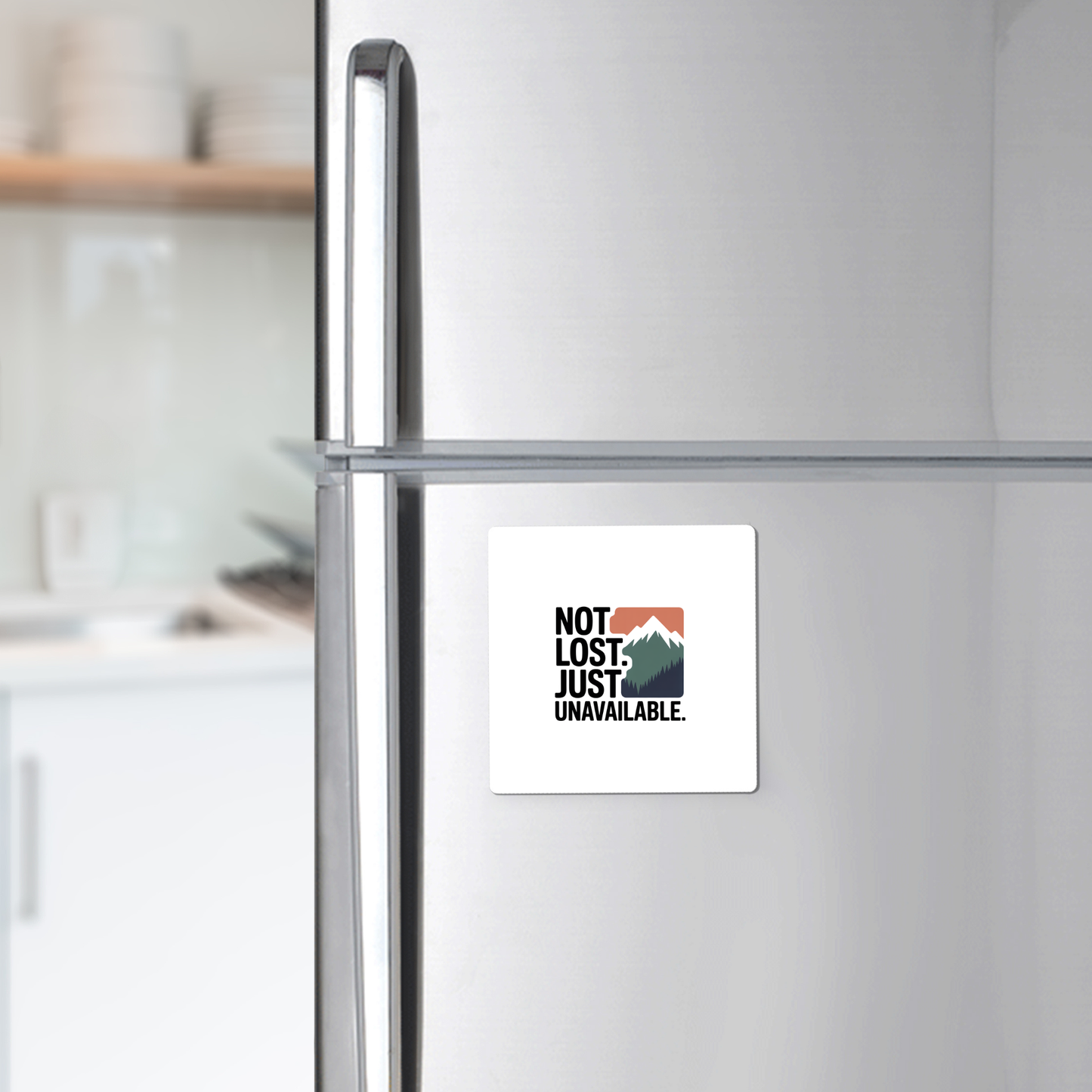 The Not Lost Just Unavailable Adventure Magnet by Mirabilia Boutique features a mountain graphic and the phrase "NOT LOST. JUST UNAVAILABLE."—a unique outdoor gift for adventurers, shown on a stainless steel fridge with a blurred kitchen in the background.