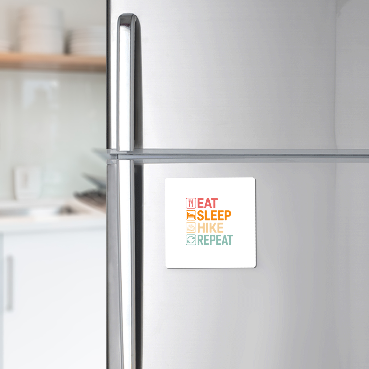 A stainless steel refrigerator features the "Eat Sleep Hike Repeat Magnet" by Mirabilia Boutique, with colorful bold letters. The softly blurred kitchen background makes this a perfect hiking gift for outdoor lovers.