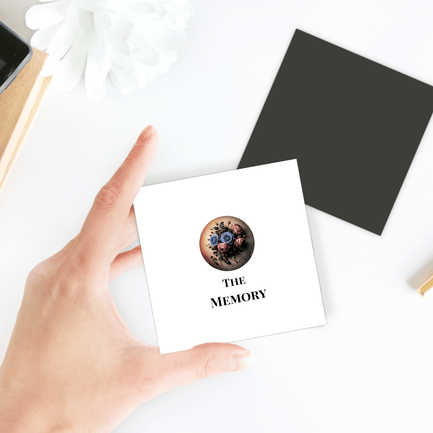 A hand holds "The Memory | Dead Regency Magnet" by Mirabilia Boutique, featuring a round gothic floral design. Nearby are a black card, white flower, and part of a gold and black object on a white surface.