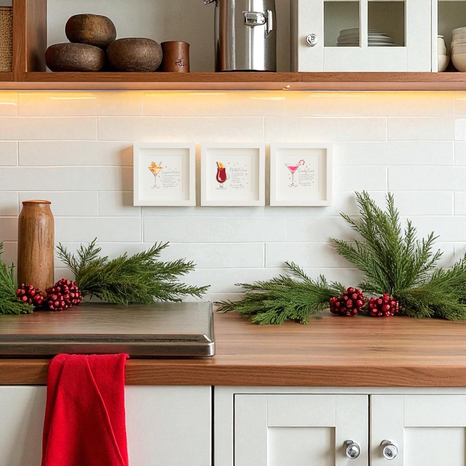 A kitchen with white cabinets and a wooden countertop features Mirabilia Boutique’s Classic Christmas Cocktails 5x5 Shadowbox Trio holiday bar art above the backsplash, evergreen decor, red berries, and a red towel in the foreground.