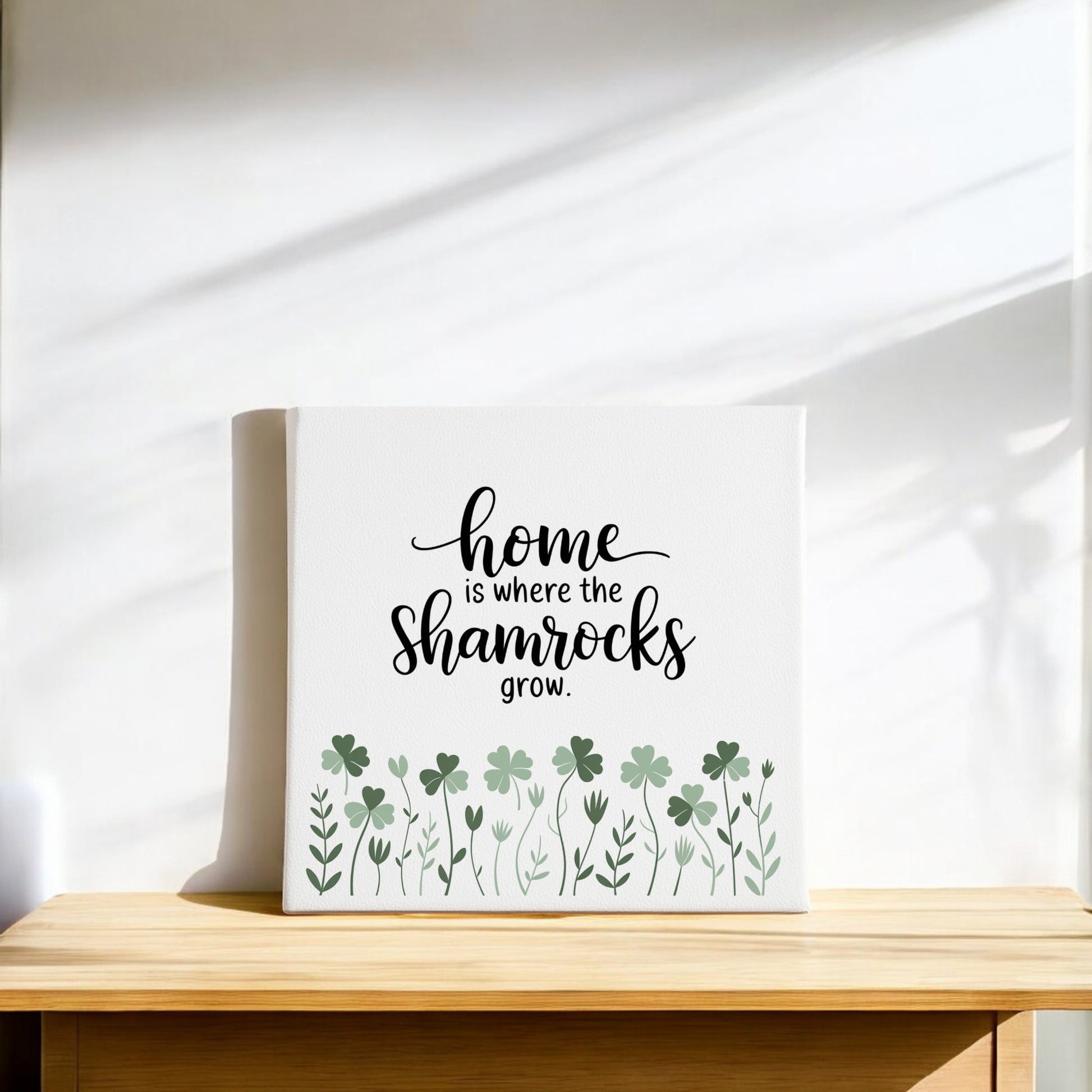 The "Home Is Where the Shamrocks Grow" Canvas by Mirabilia Boutique features green shamrocks on a white background, making it ideal Irish home decor or a thoughtful St. Patrick's Day gift. Displayed on light wood with sunlight in the background.