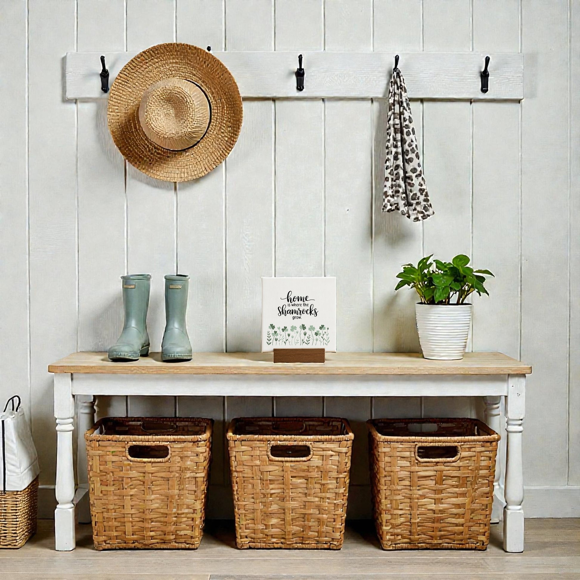 A tidy entryway features a bench with three woven baskets, a potted plant, and the Mirabilia Boutique "Home Is Where the Shamrocks Grow" mini sign. Above are hooks with a straw hat and scarf. Green rain boots sit on the bench, with another basket on the floor nearby.