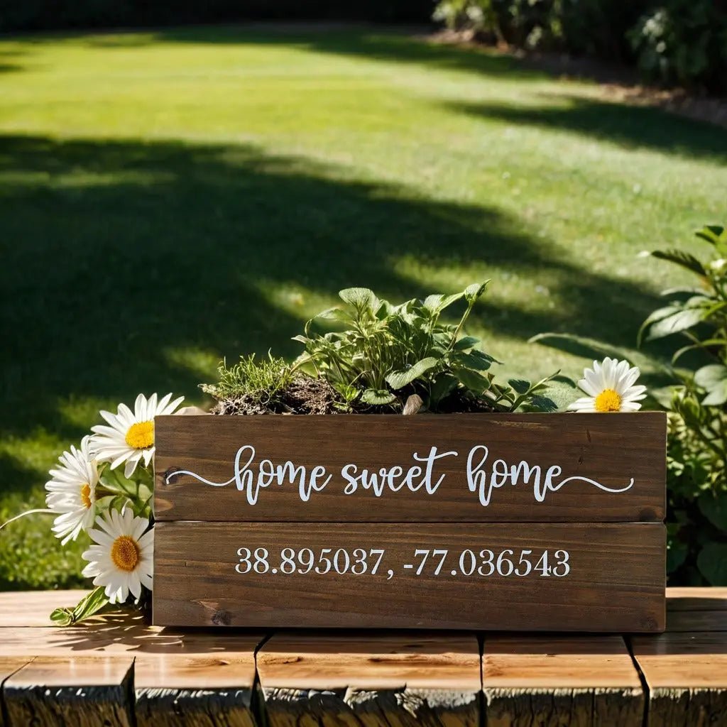 A Mirabilia Boutique's charming "Home Sweet Home" wall plaque enhances a wooden planter box filled with daisies and green plants, resting on a bench with the customizable coordinates "38.895037, -77.036543," against a backdrop of lush green grass and a vibrant garden.
