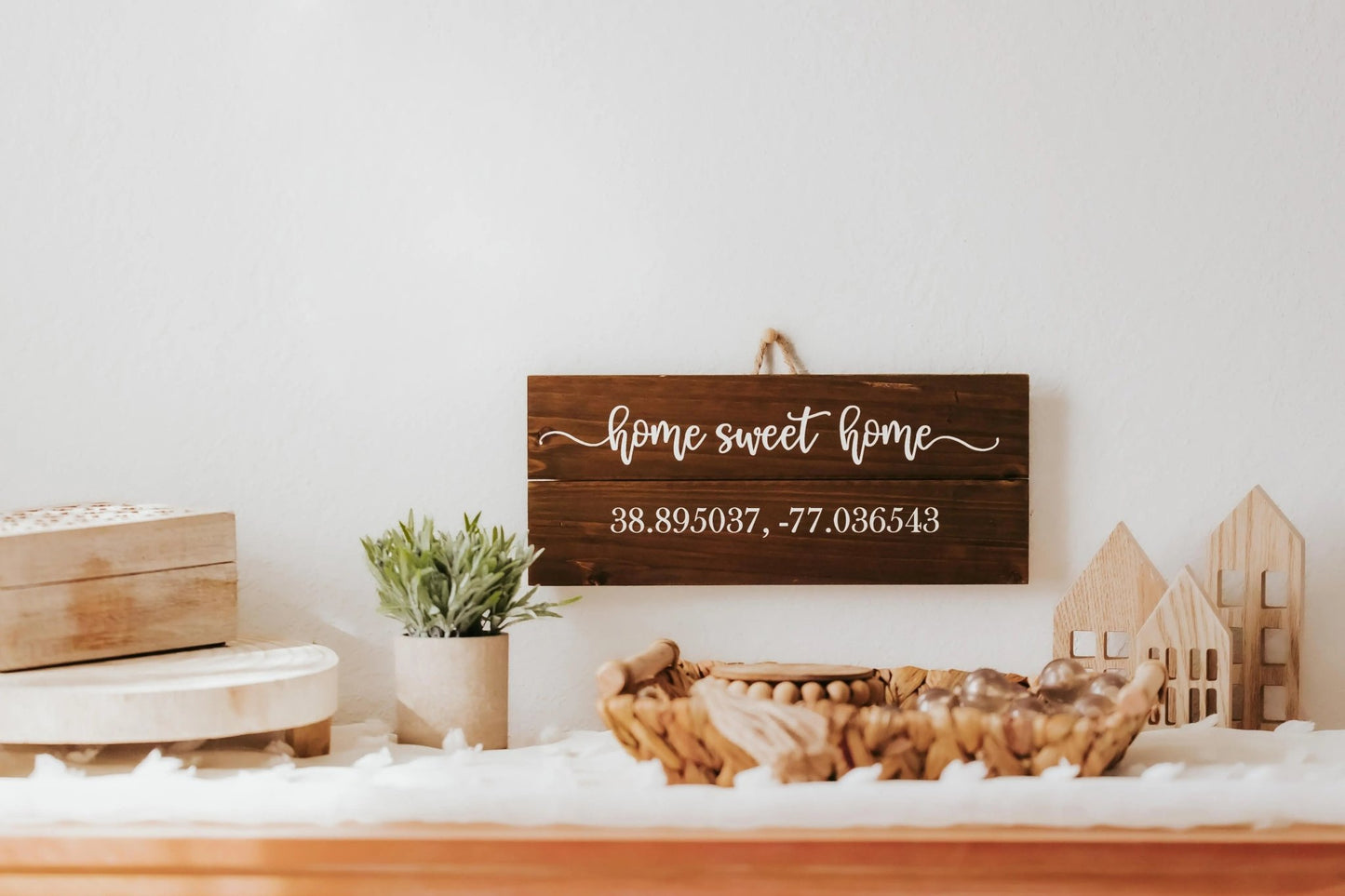 A charming wall plaque from Mirabilia Boutique, the "Charming Home Sweet Home Wall Plaque for Cozy Rustic Decor," showcases the phrase "Home Sweet Home" with customizable coordinates below. It hangs above a shelf decorated with a plant, small wooden houses, and two baskets filled with items.