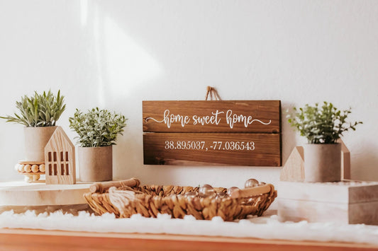 A charming "Home Sweet Home" wall plaque from Mirabilia Boutique adds a touch of rustic elegance with customizable coordinates. Below, a shelf displays potted plants, a quaint wooden house, a basket filled with decorative stones, and candles resting on a fabric surface.