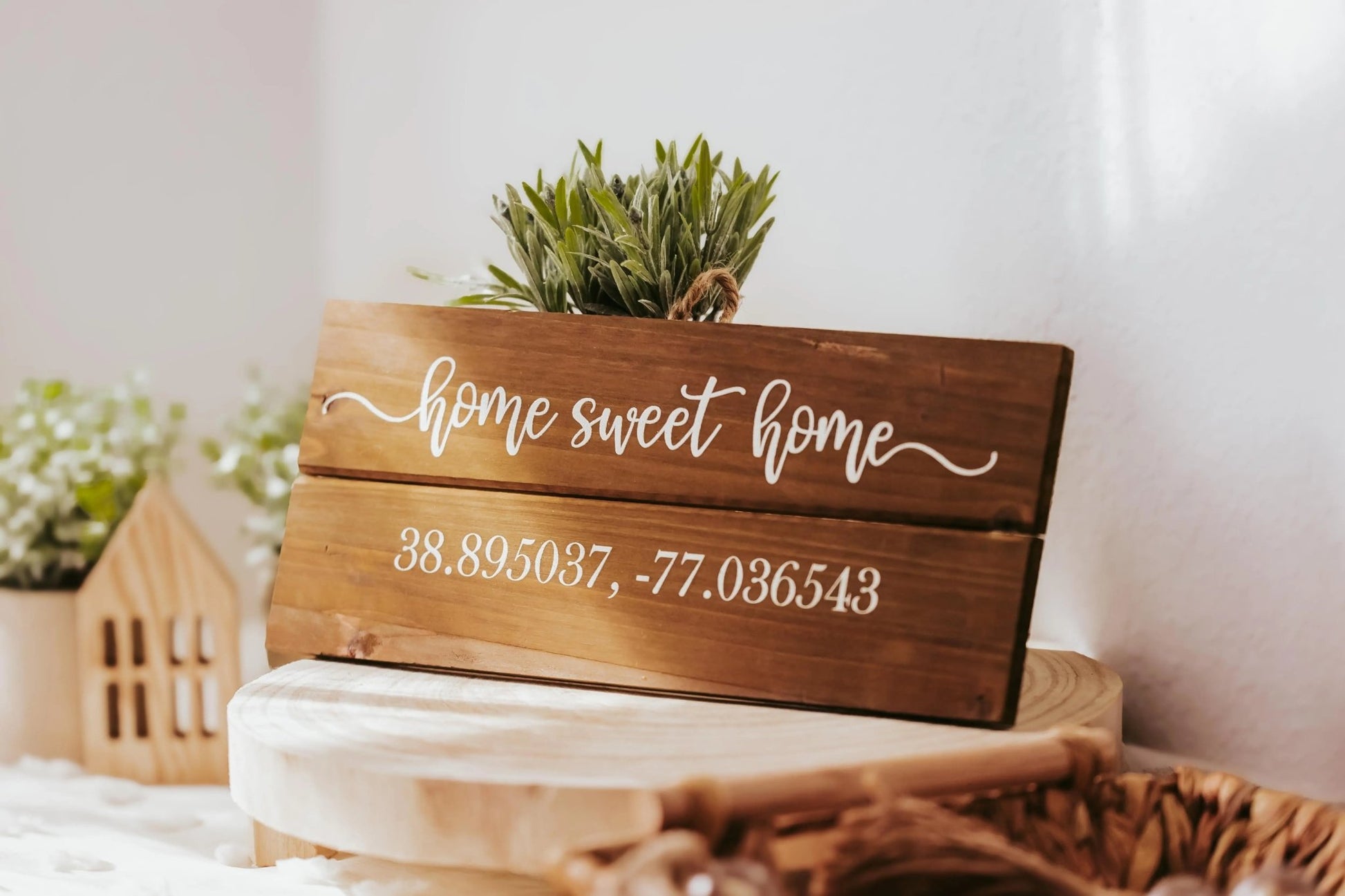 The Mirabilia Boutique’s Charming Home Sweet Home Wall Plaque for Cozy Rustic Decor features elegant white script with "Home Sweet Home" and customizable coordinates "38.895037, -77.036543" below. It is beautifully enhanced with small green plants and decorative wooden house models, all displayed on a light-colored surface.