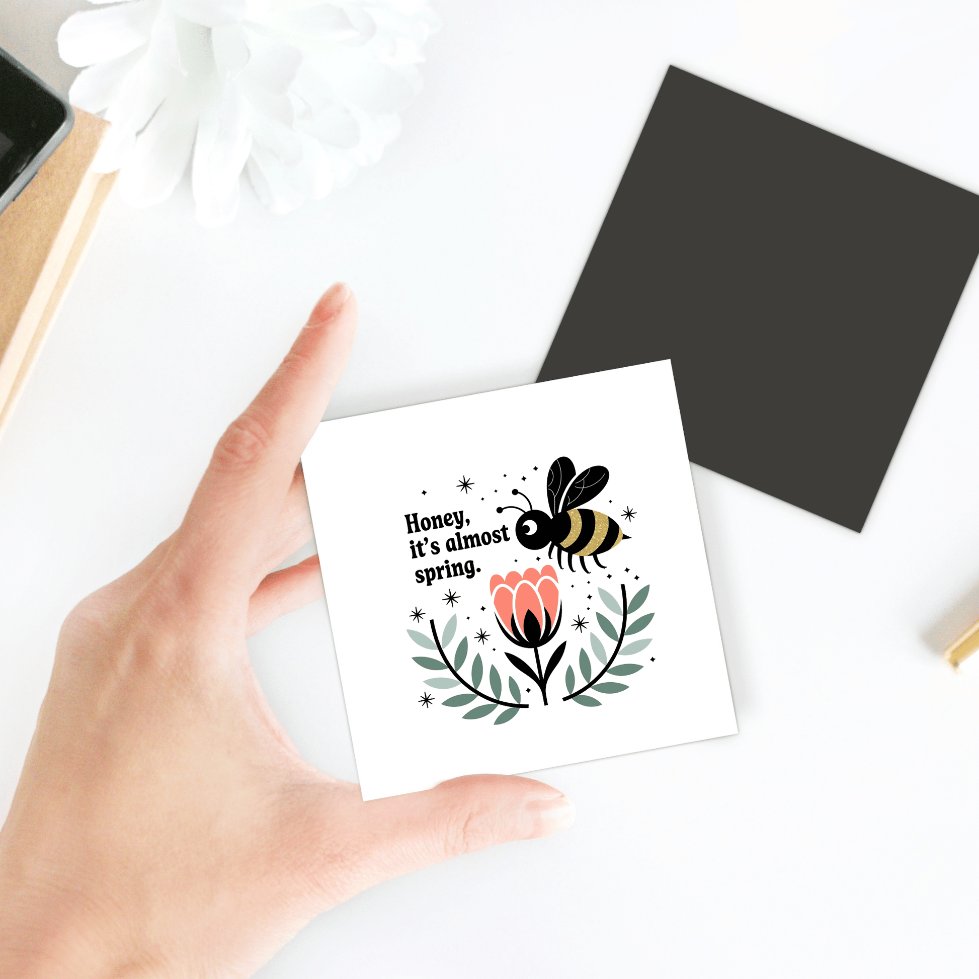 A hand holds the "Honey It’s Almost Spring Magnet" by Mirabilia Boutique, featuring a bee above a pink flower and green leaves—perfect as cute spring decor for your fridge. A black square and white flower rest on the white tabletop.