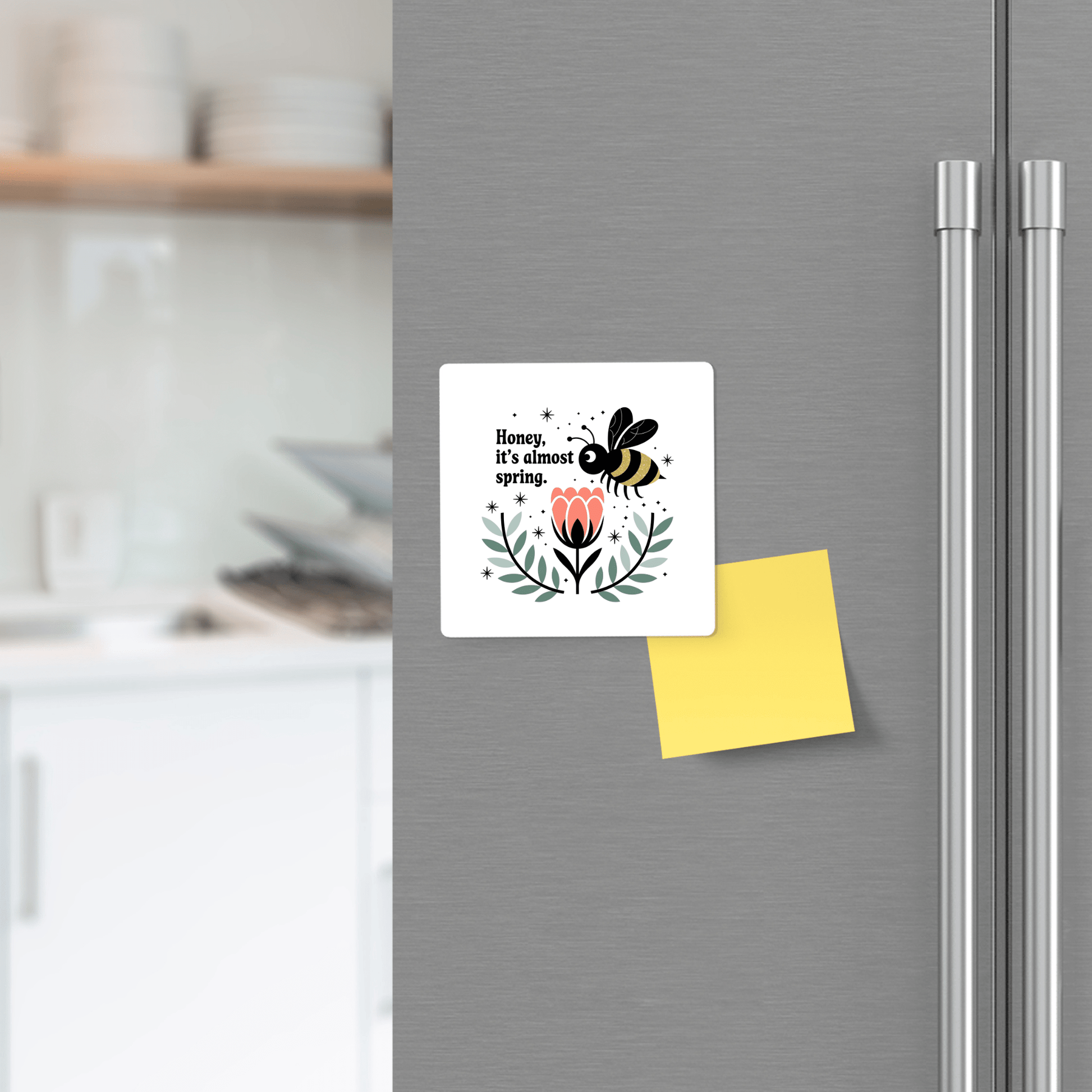 The Honey It's Almost Spring Magnet by Mirabilia Boutique is a cute bee fridge magnet with a pink flower and cheerful spring message, perfect for brightening up your stainless steel refrigerator. A yellow sticky note sits just below it.