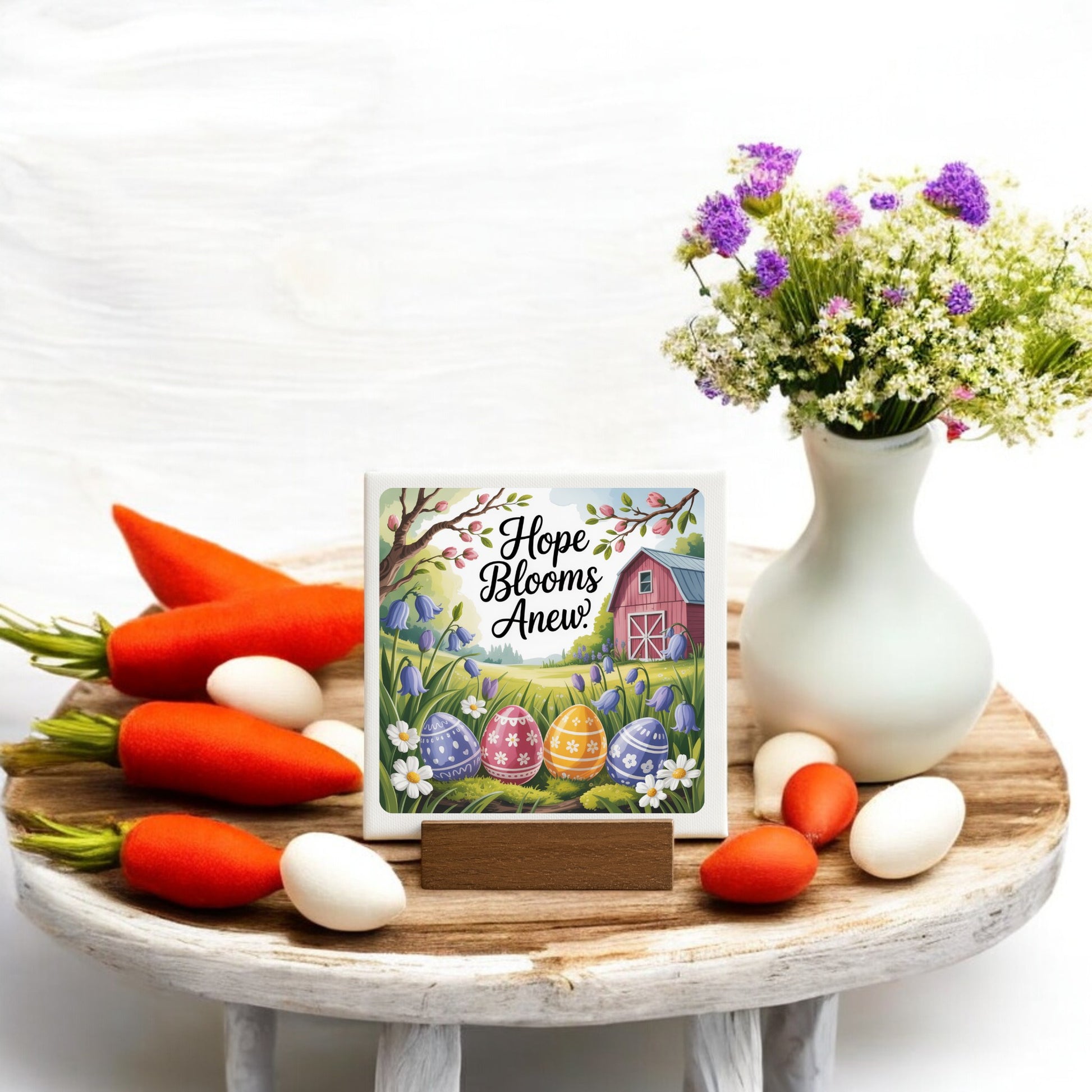 The Hope Blooms Anew Mini Canvas – 4x4 Spring Sign by Mirabilia Boutique, featuring Easter eggs, flowers, and a red barn, is displayed on a tray with carrots, eggs, Easter decor, and a white vase of purple and white flowers.