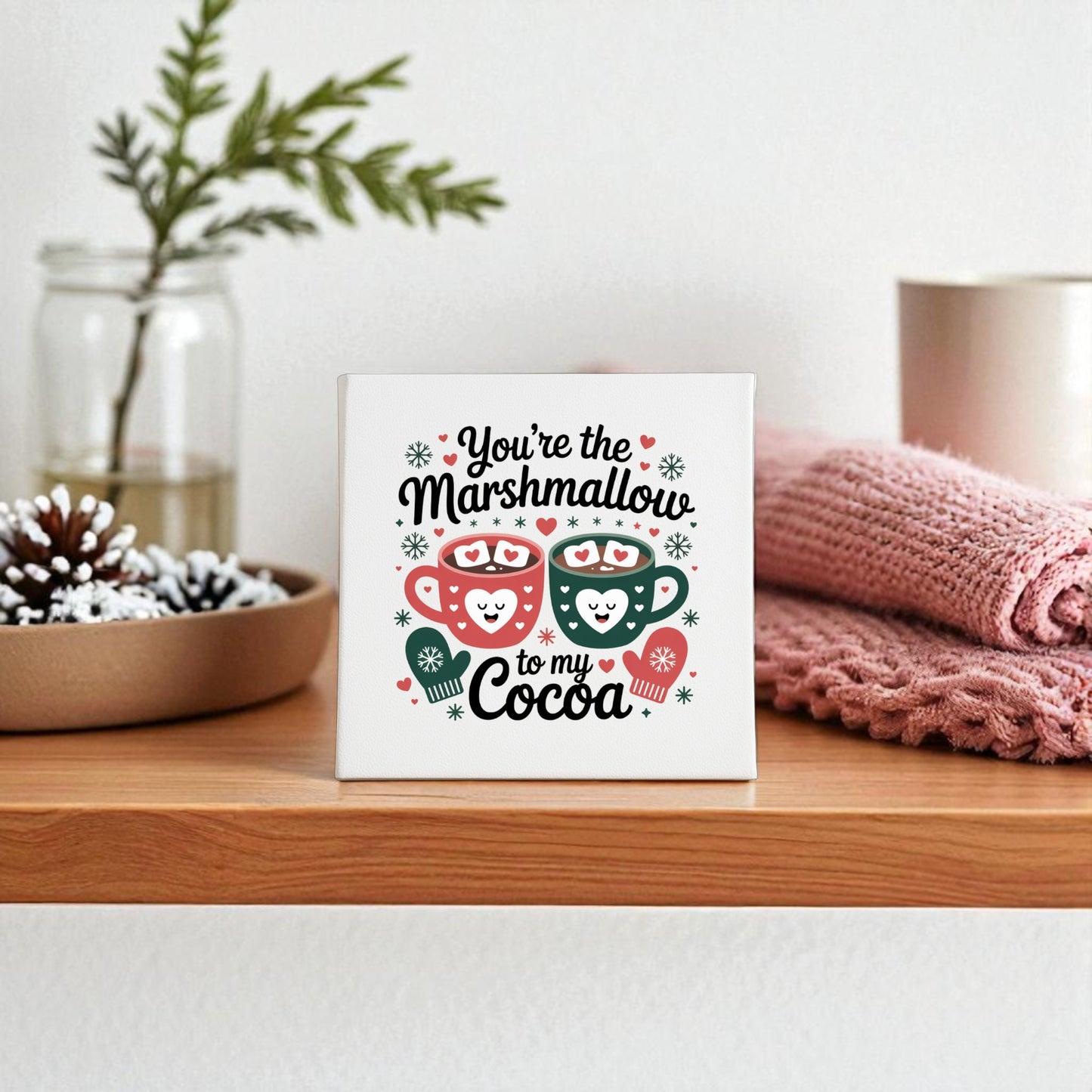 The Mirabilia Boutique "Marshmallow to My Cocoa" 8x8 Winter Canvas Art adds cozy charm to your kitchen shelf, paired with mugs, marshmallows, pink cloth, a pinecone, and greenery in a glass jar for festive winter decor.