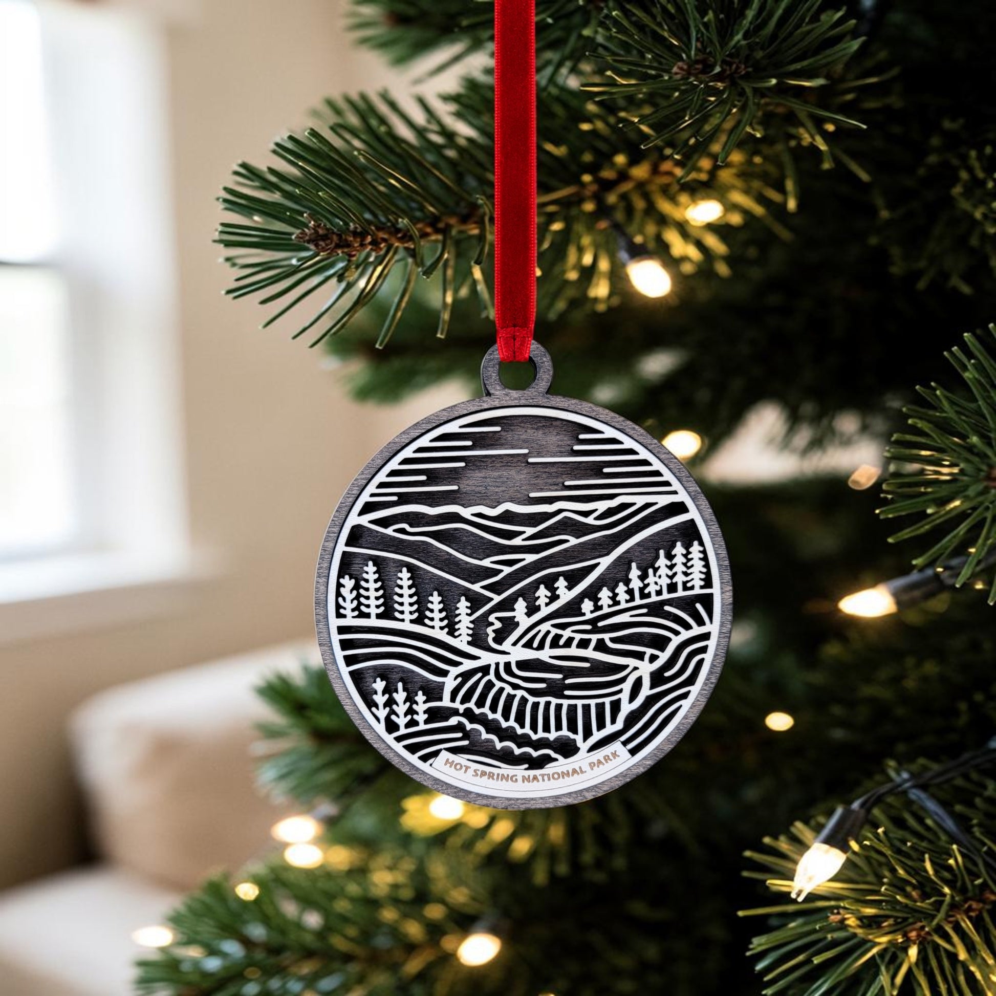 The Hot Springs National Park Line Art Christmas Ornament by Mirabilia Boutique hangs on a Christmas tree adorned with lights. This handcrafted wooden oval ornament features an intricate landscape design depicting mountains, trees, and a river, along with the inscription “Hot Springs National Park” that beautifully captures the spirit of Bathhouse Row. A window appears softly blurred in the background.