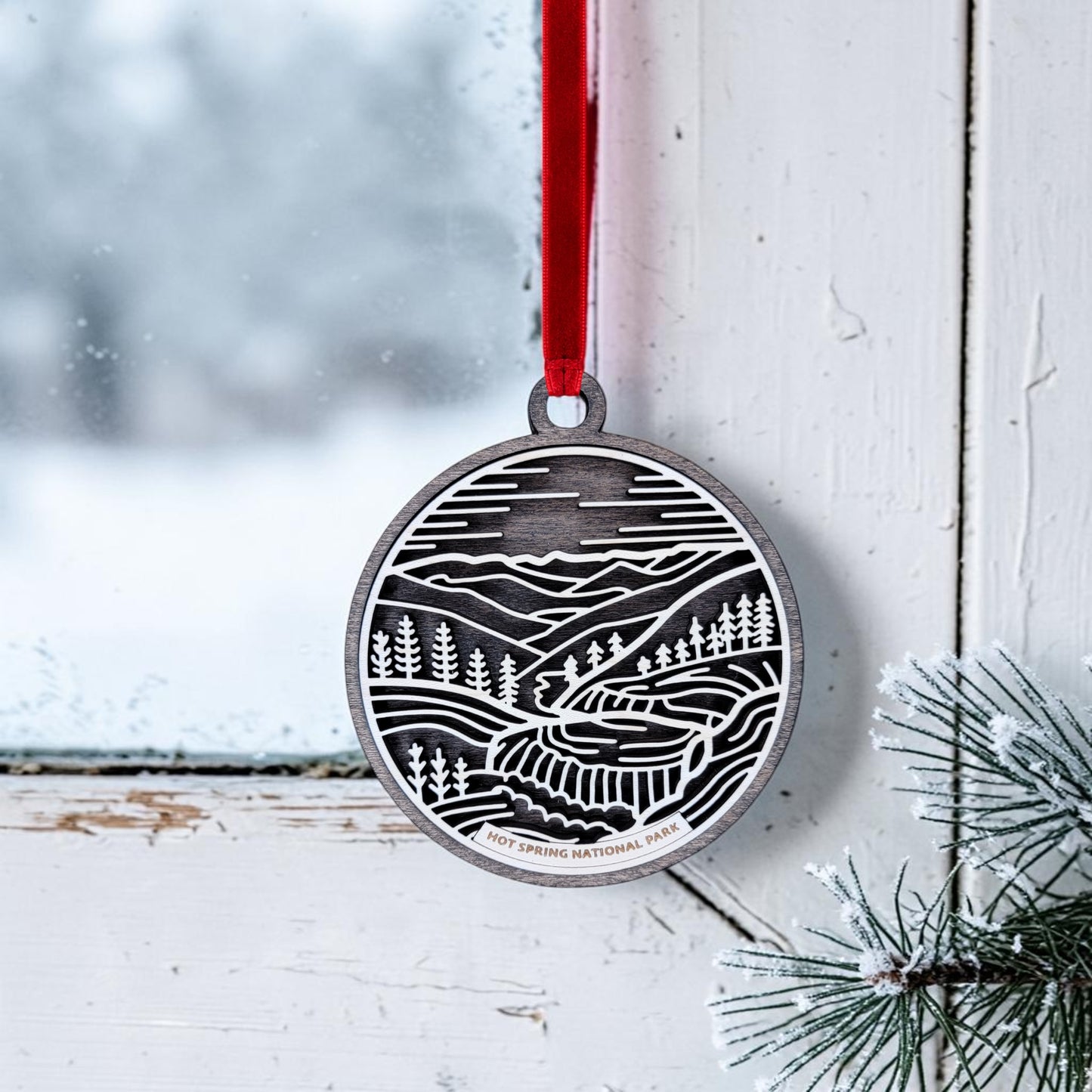 A charming keepsake from Mirabilia Boutique, the Hot Springs National Park Line Art Christmas Ornament features an engraved landscape of mountains, trees, and a river. This metallic ornament hangs from a red ribbon on a snowy window, with pine needles tucked into the corner.