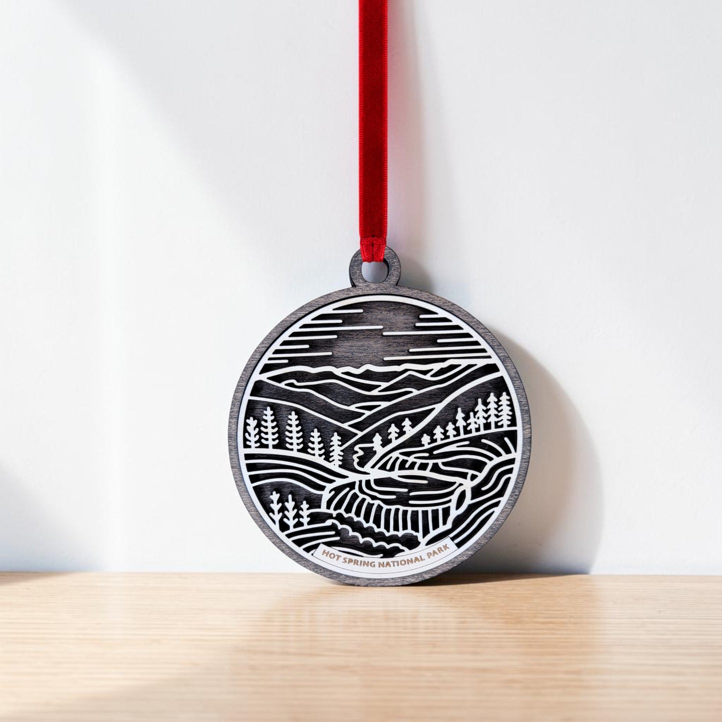 The Hot Springs National Park Line Art Christmas Ornament by Mirabilia Boutique is a handcrafted wooden round medal, adorned with a black and white landscape illustration of mountains, trees, and a winding path. It hangs from a red ribbon against a white wall and wooden surface, beautifully capturing the essence of Hot Springs National Park.