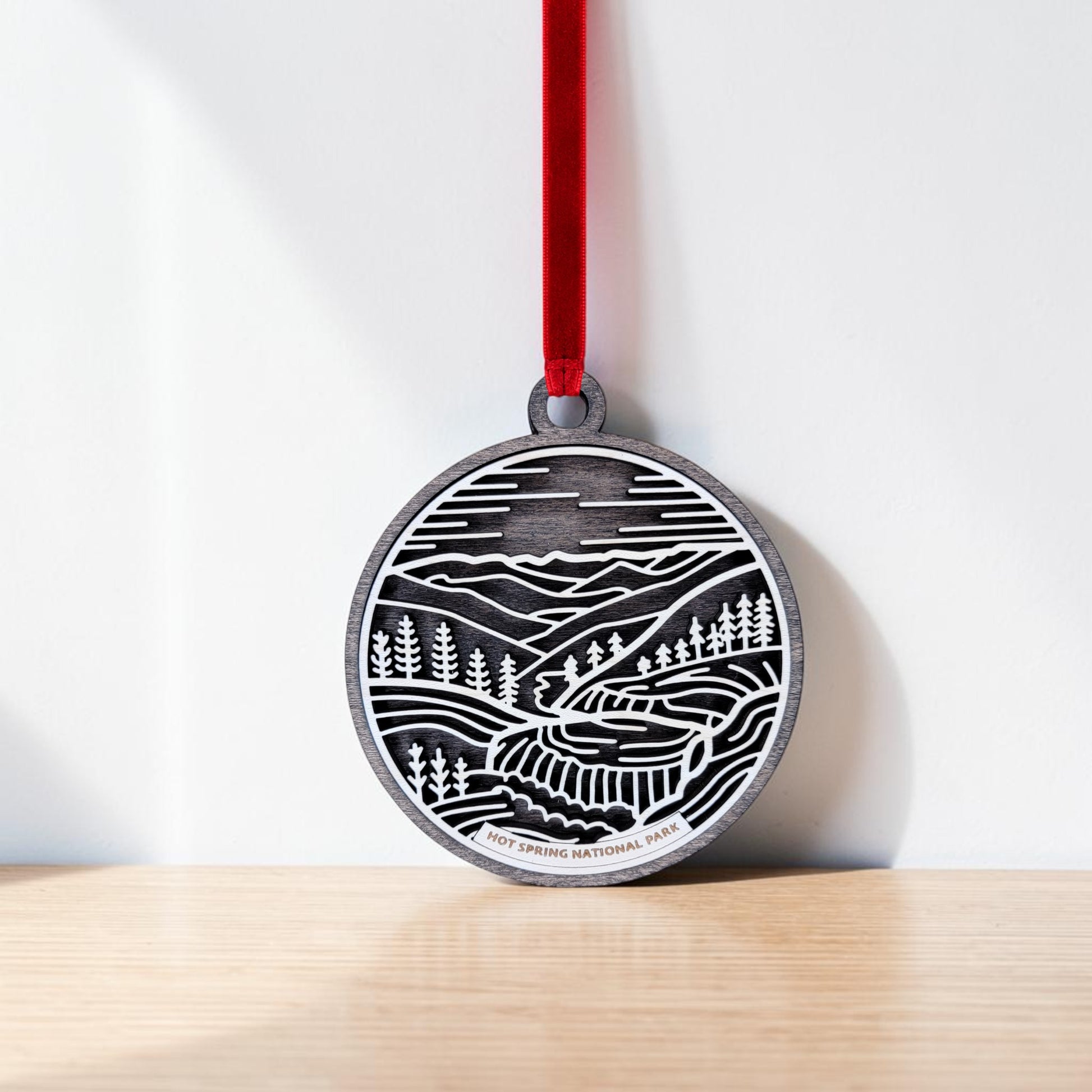 The Hot Springs National Park Line Art Christmas Ornament by Mirabilia Boutique is a handcrafted wooden round medal, adorned with a black and white landscape illustration of mountains, trees, and a winding path. It hangs from a red ribbon against a white wall and wooden surface, beautifully capturing the essence of Hot Springs National Park.