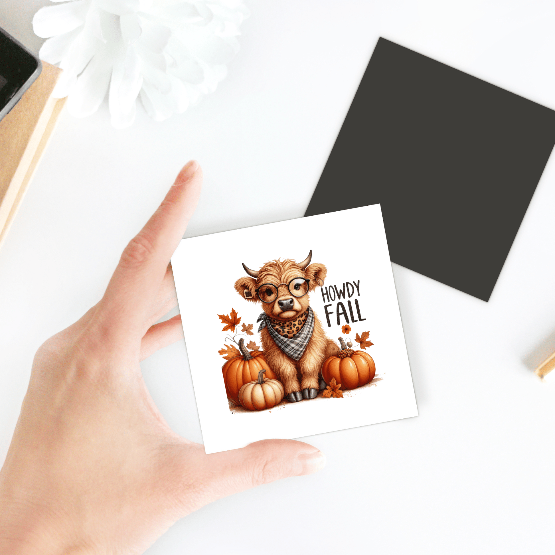 A hand holds the Howdy Fall Highland Cow Magnet by Mirabilia Boutique, showing a cute cow in glasses and scarf among pumpkins and autumn leaves—adorable autumn kitchen decor. Nearby are a white flower and black square on a white surface.