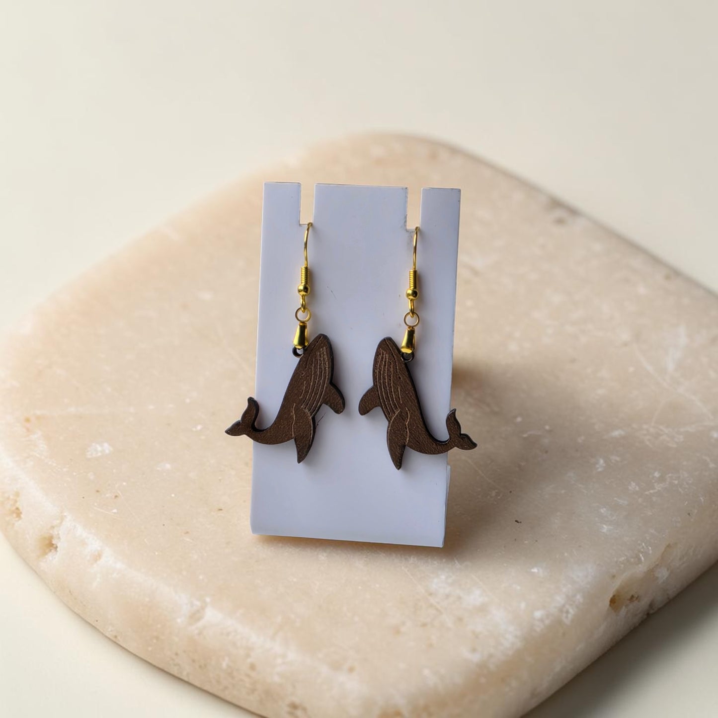 Humpback Whale Earrings by Mirabilia Boutique—lasercut from walnut wood—displayed on a white stand atop a beige stone, perfect ocean-inspired jewelry for marine life lovers.