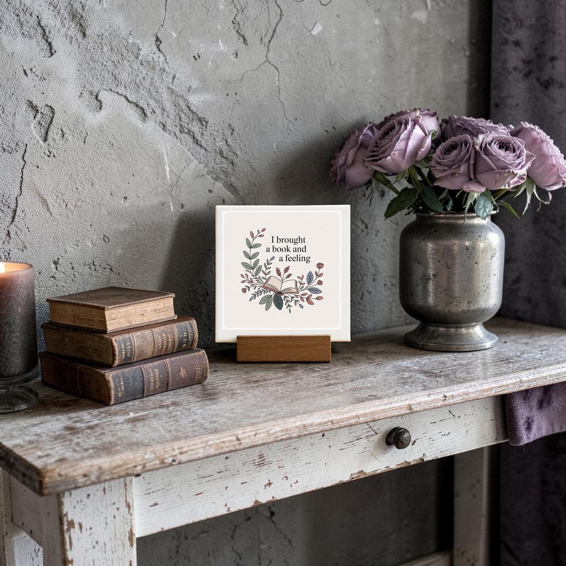 A rustic table displays vintage books, a lit candle, purple flowers in a vase, and the Mirabilia Boutique "I Brought a Book and a Feeling" mini sign—perfect reading decor for any book lover’s gift.
