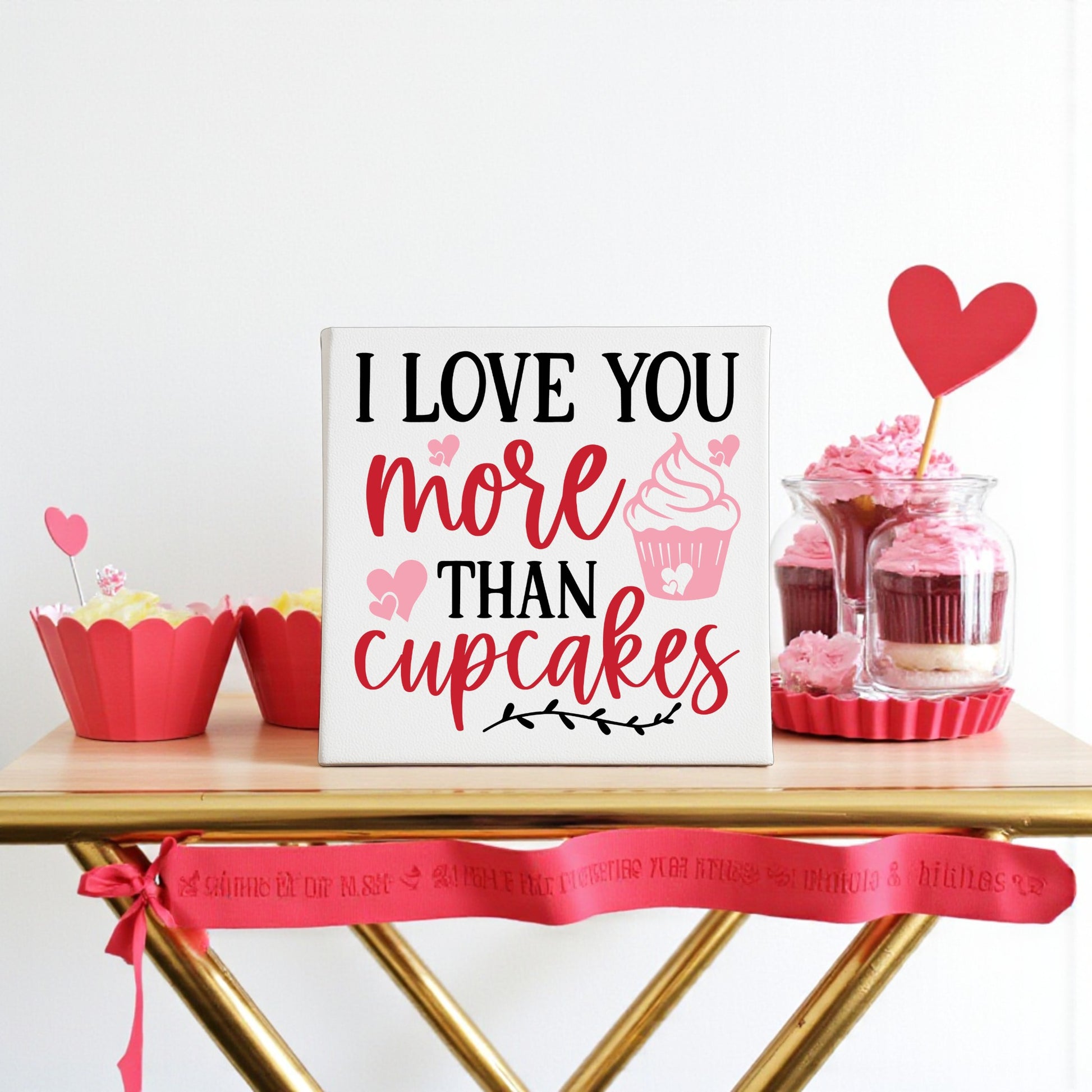 An 8x8 "I Love You More Than Cupcakes" canvas sign by Mirabilia Boutique sits on a table surrounded by cupcakes, heart-topped jars, and red ribbon, creating a charming romantic Valentine’s decor.