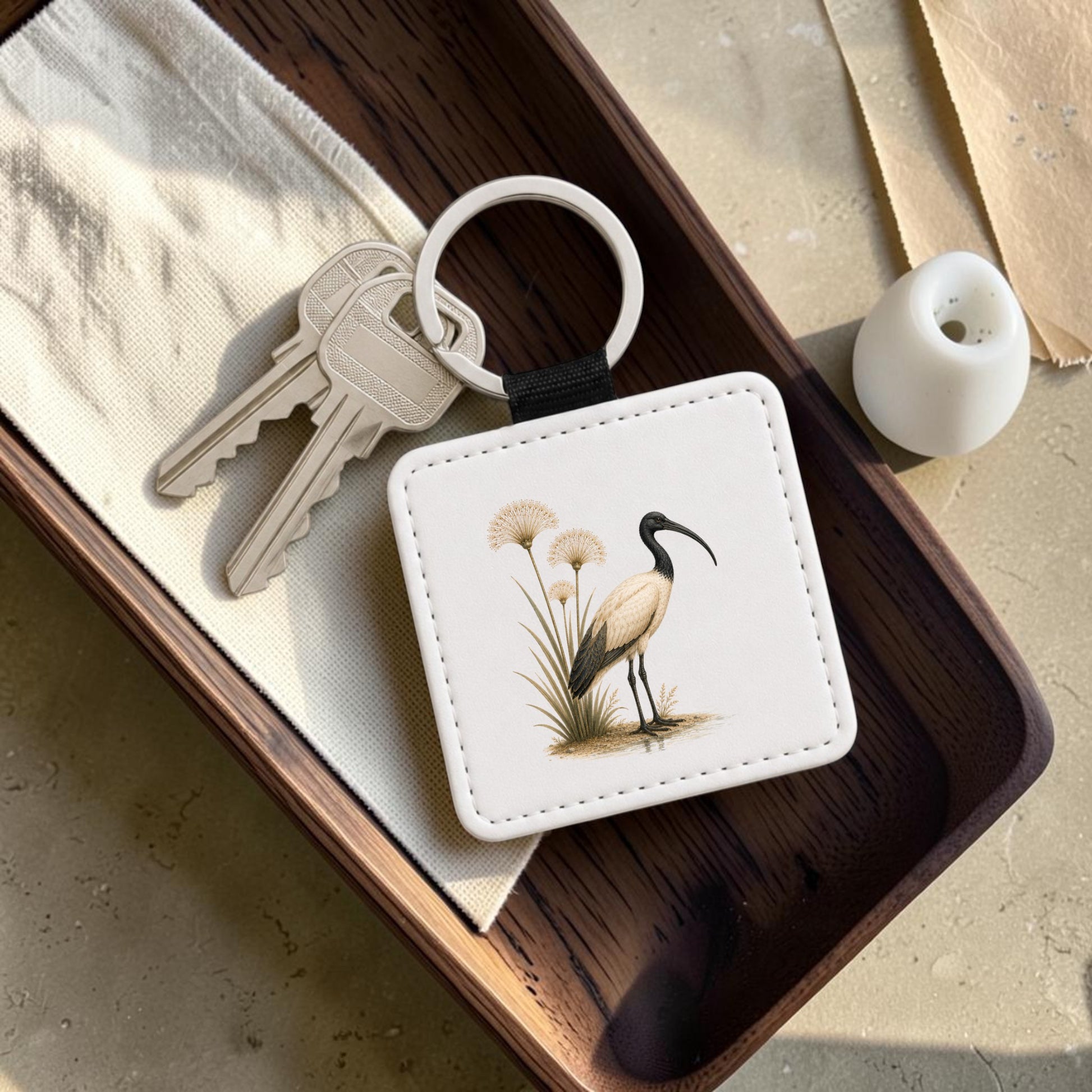 The Ibis & Papyrus Keychain | Ancient Egyptian Art by Mirabilia Boutique, a square keychain adorned with an illustration of an ibis and papyrus, rests in a wooden tray next to two silver keys, a folded white napkin, and a small white vase on a beige surface.