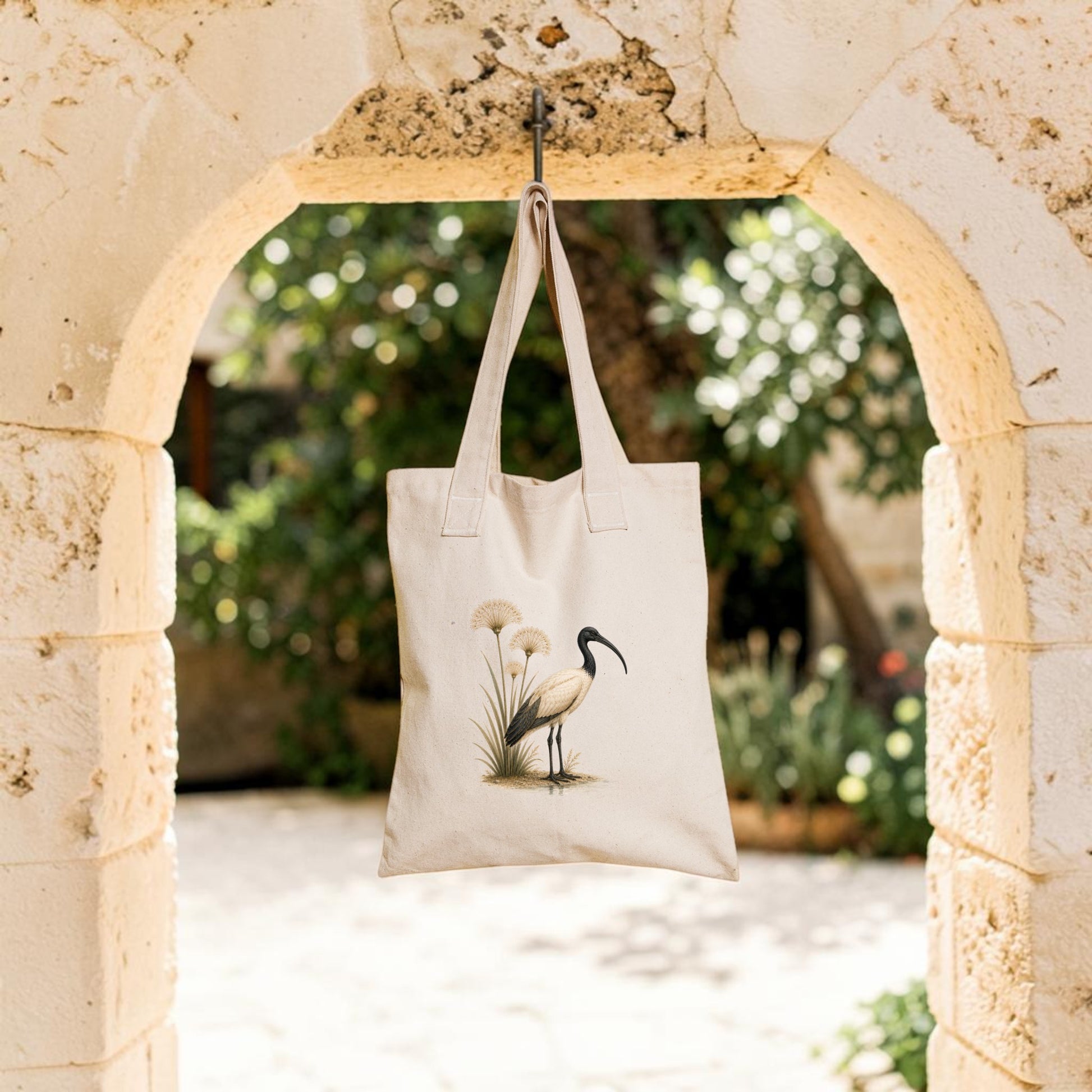 A beige Ibis & Papyrus Tote Bag from Mirabilia Boutique, featuring ancient Egyptian art of an ibis and papyrus plants, hangs on a stone archway with a sunlit garden softly blurred in the background.