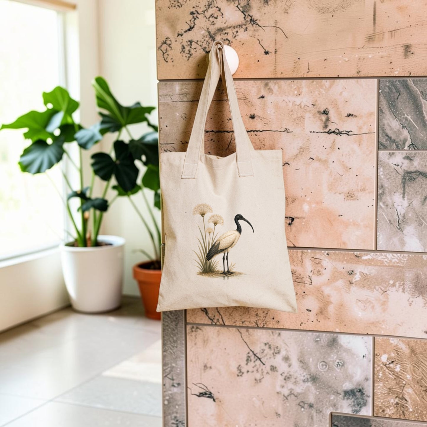 The “Ibis & Papyrus Tote Bag | Ancient Egyptian Art” by Mirabilia Boutique, featuring a heron and papyrus illustration in beige, hangs on a textured wall; green potted plants sit in the background by a sunlit window.