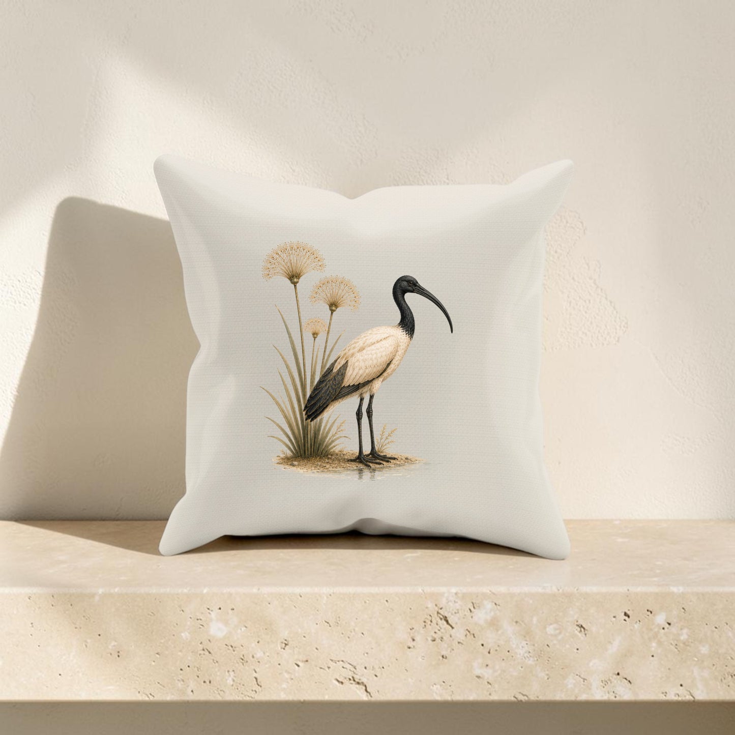 The Ibis & Papyrus Pillow from Mirabilia Boutique features a graceful illustration of an ibis standing beside tall grass, set against a white background. Placed on a beige stone or concrete shelf with a textured light-colored wall, this decorative accent echoes the elegance of Egyptian home decor and lends a timeless charm to any space.