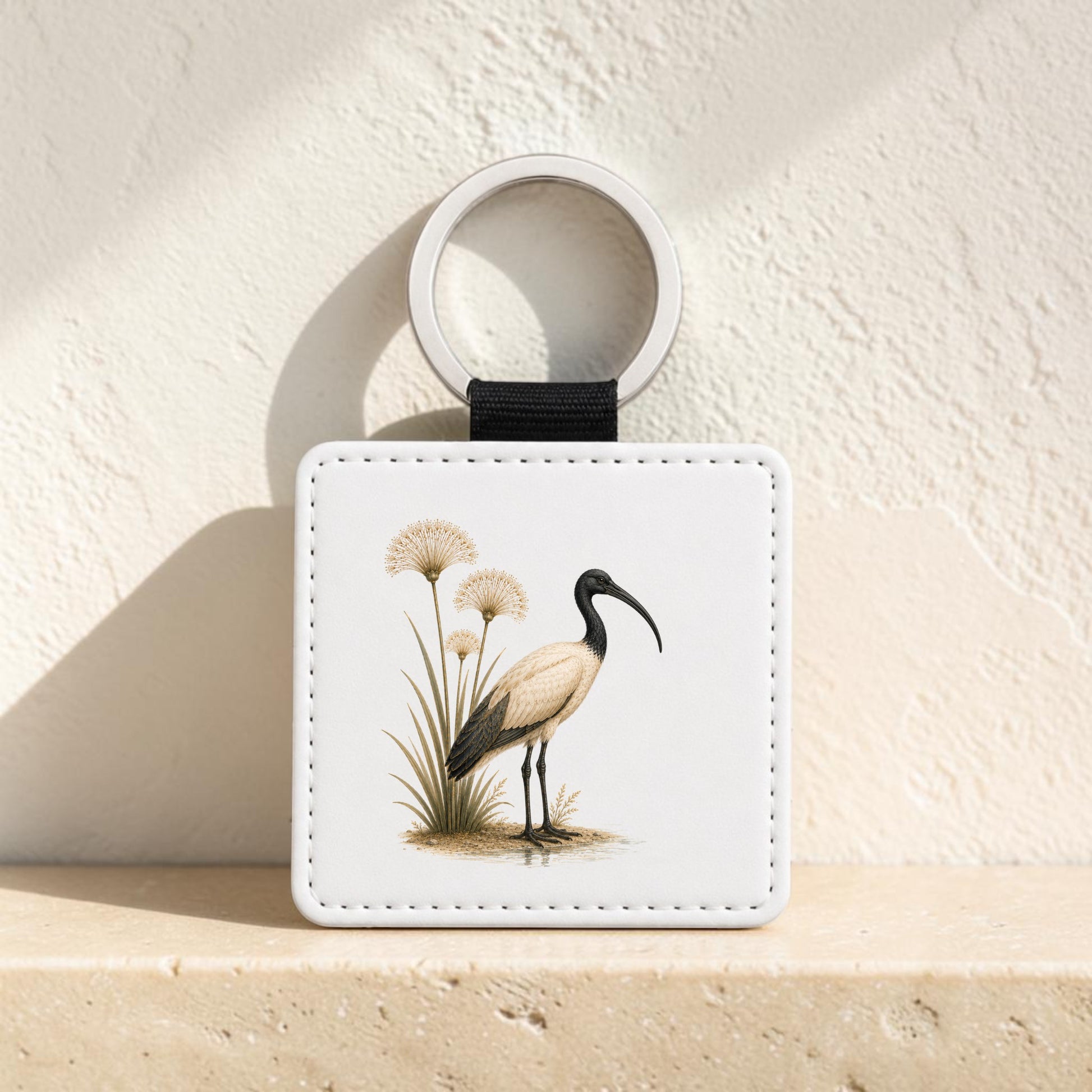 The Ibis & Papyrus Keychain | Ancient Egyptian Art by Mirabilia Boutique is a white square keychain with a black strap, adorned with an illustration of an ibis and papyrus reminiscent of ancient Egypt. The design depicts a wading bird with a long beak standing among grasses. The keychain is shown upright on a beige stone surface against a textured wall.