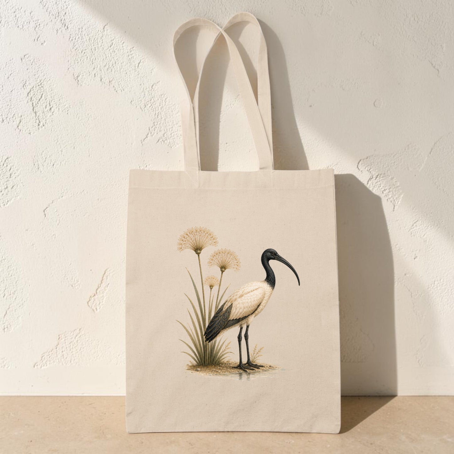 The Ibis & Papyrus Tote Bag from Mirabilia Boutique showcases ancient Egyptian art, with a beige landscape design featuring an illustration of a black and white ibis standing beside tall papyrus grasses, set against a textured light-colored wall.
