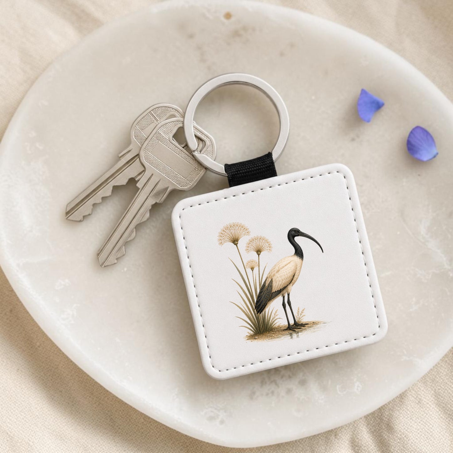On a white dish, the Ibis & Papyrus Keychain from Mirabilia Boutique—featuring an enchanting bird and grass illustration inspired by ancient Egyptian art—rests beside two silver keys and two delicate purple flower petals.