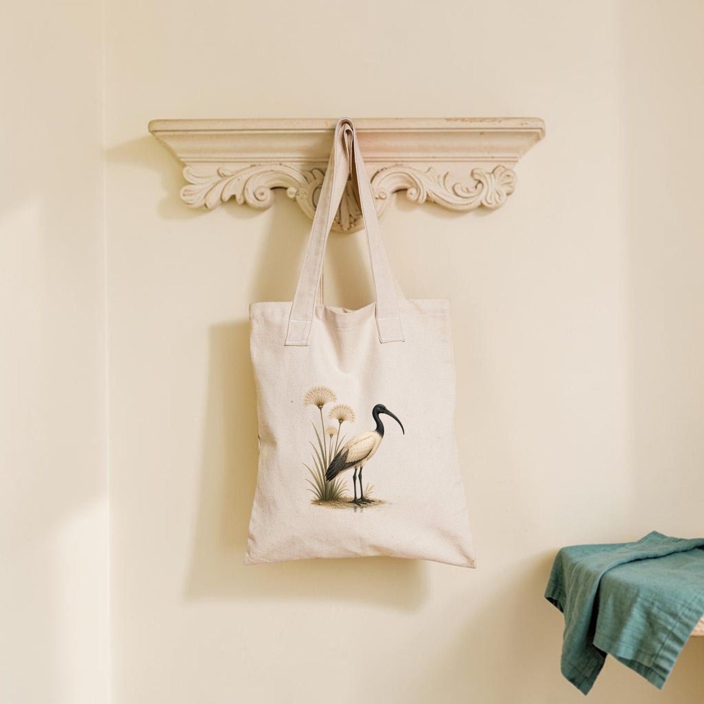 The Ibis & Papyrus Tote Bag from Mirabilia Boutique, featuring an ancient Egyptian bird and papyrus design in beige, hangs on a decorative wall shelf. A green cloth is folded on a bench to the right, set against a pale cream background wall.